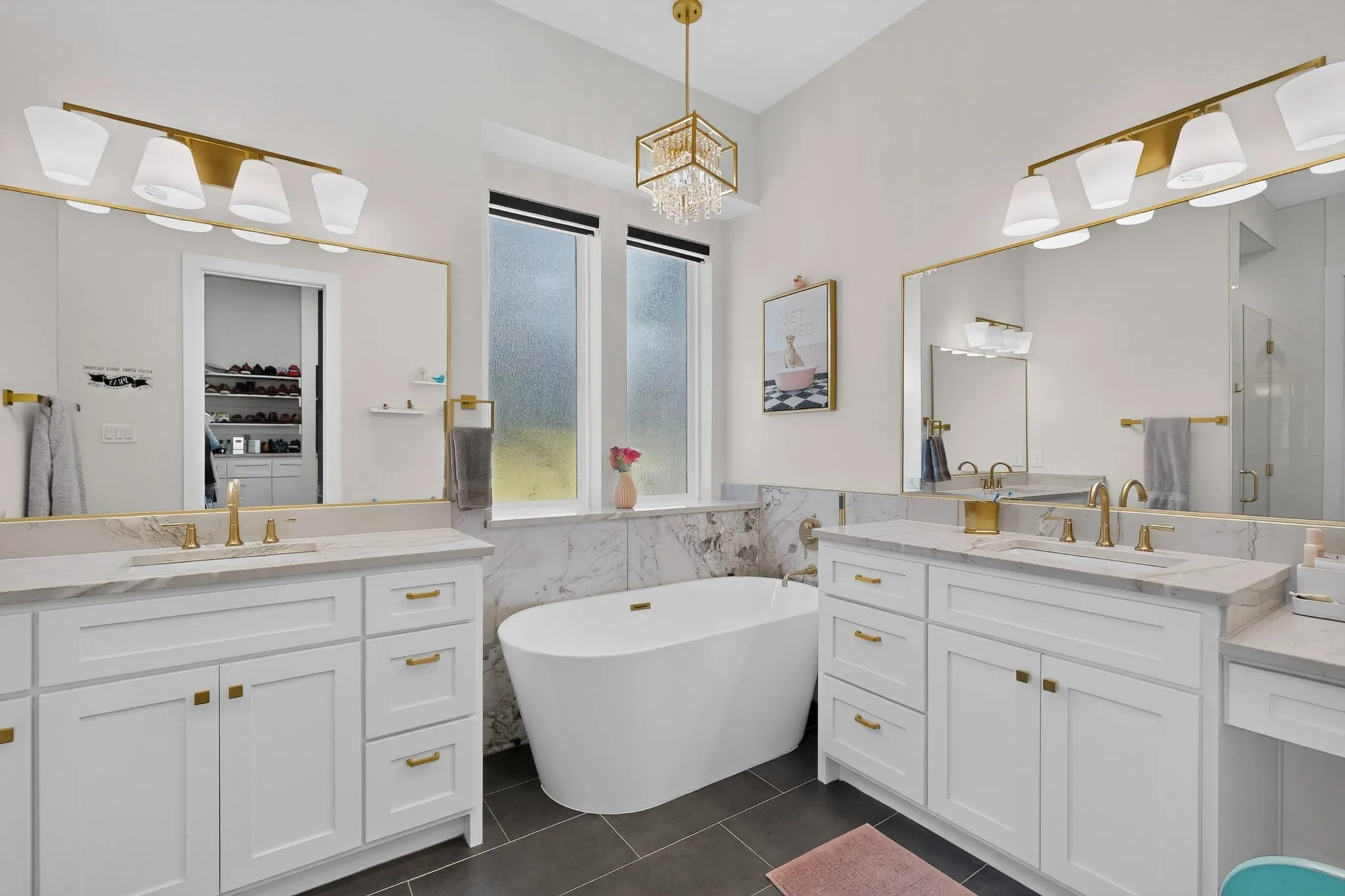Dual white vanities with gold-tone hardware, a freestanding soaking tub, large format floor tile, and gold-finish lighting fixtures