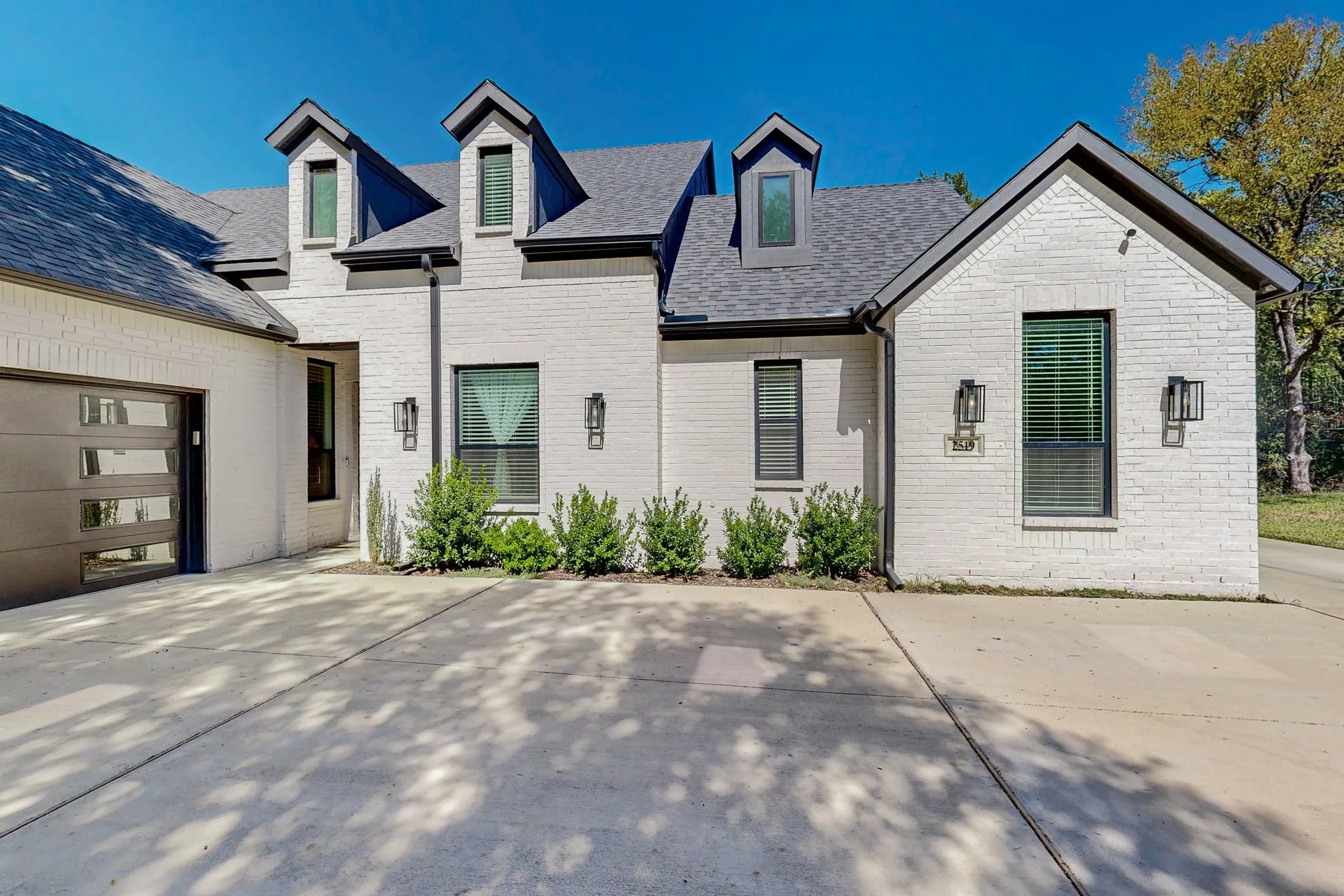 White brick exterior with a dark gray roof, prominent dormer windows, and a modern garage door featuring frosted panels