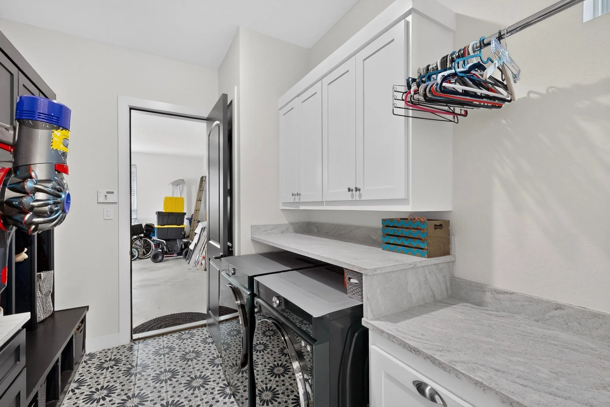 Geometric-patterned floor tile, white shaker-style cabinetry, light-toned countertop, and a clothes hanging rod