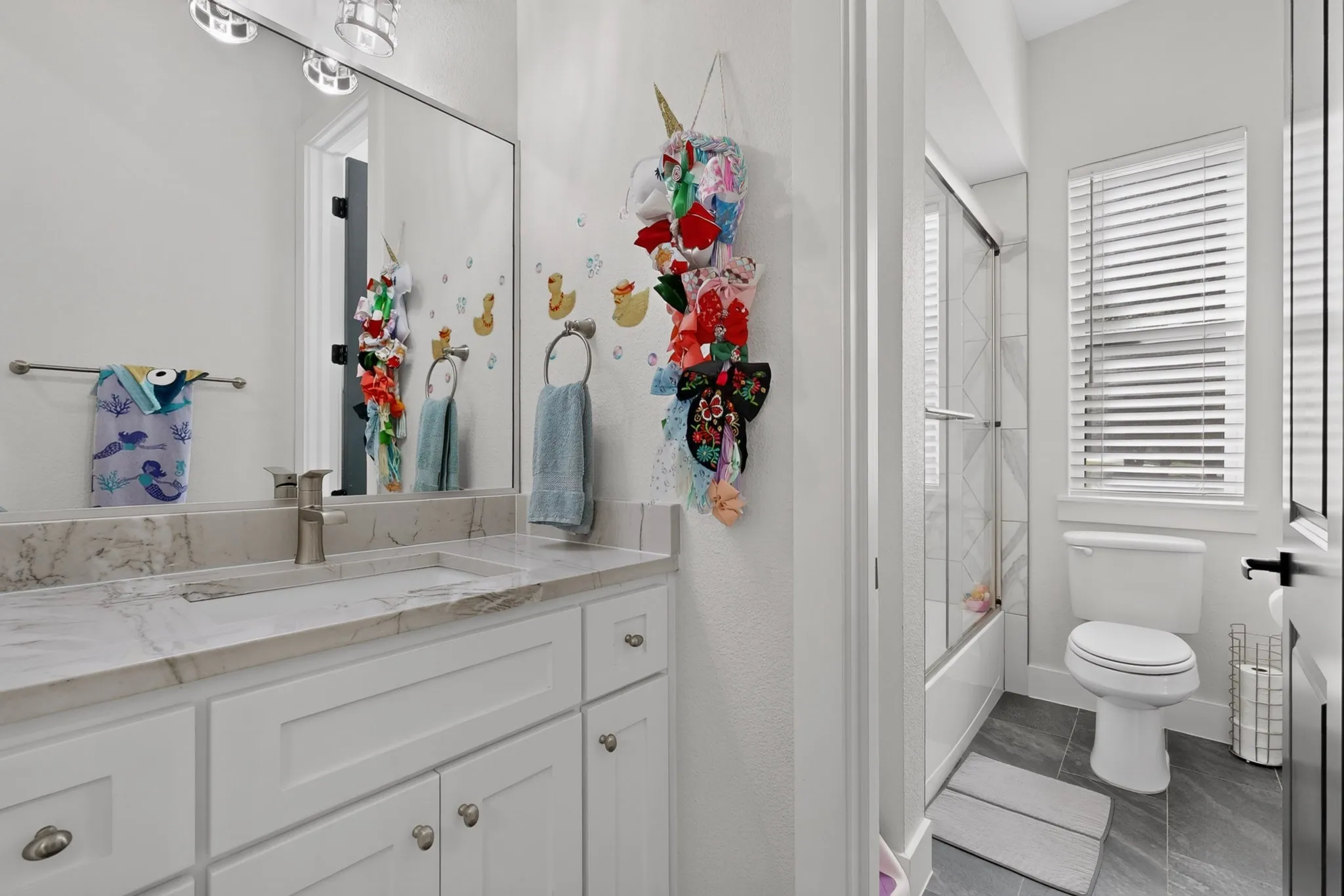 Bathroom featuring white shaker cabinetry, an undermount sink, a stone-finish countertop, and a shower-tub combination with a glass enclosure
