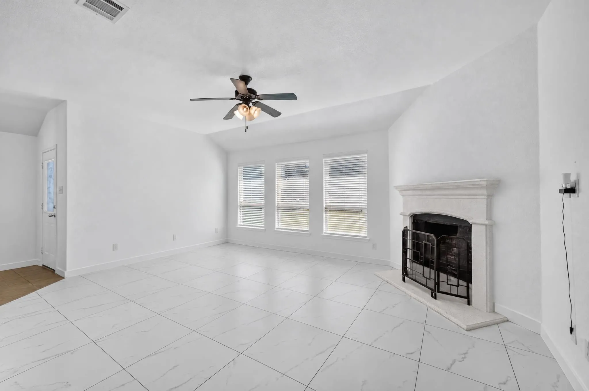 Spacious room featuring white tile flooring, a fireplace with a decorative mantel, multiple windows with blinds, and a ceiling fan with integrated lighting