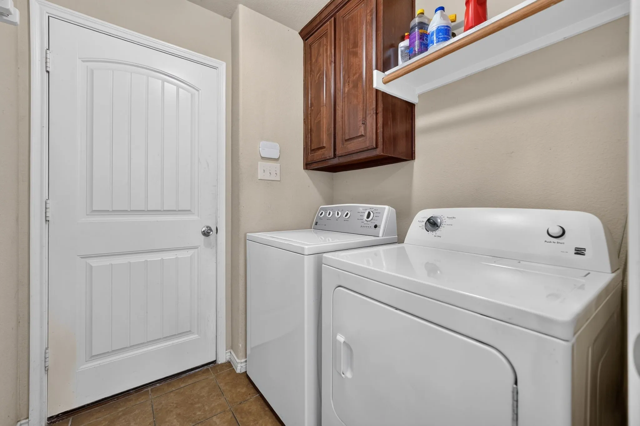 Laundry area featuring overhead wood-finish cabinetry, a wall-mounted shelving unit, and tile flooring