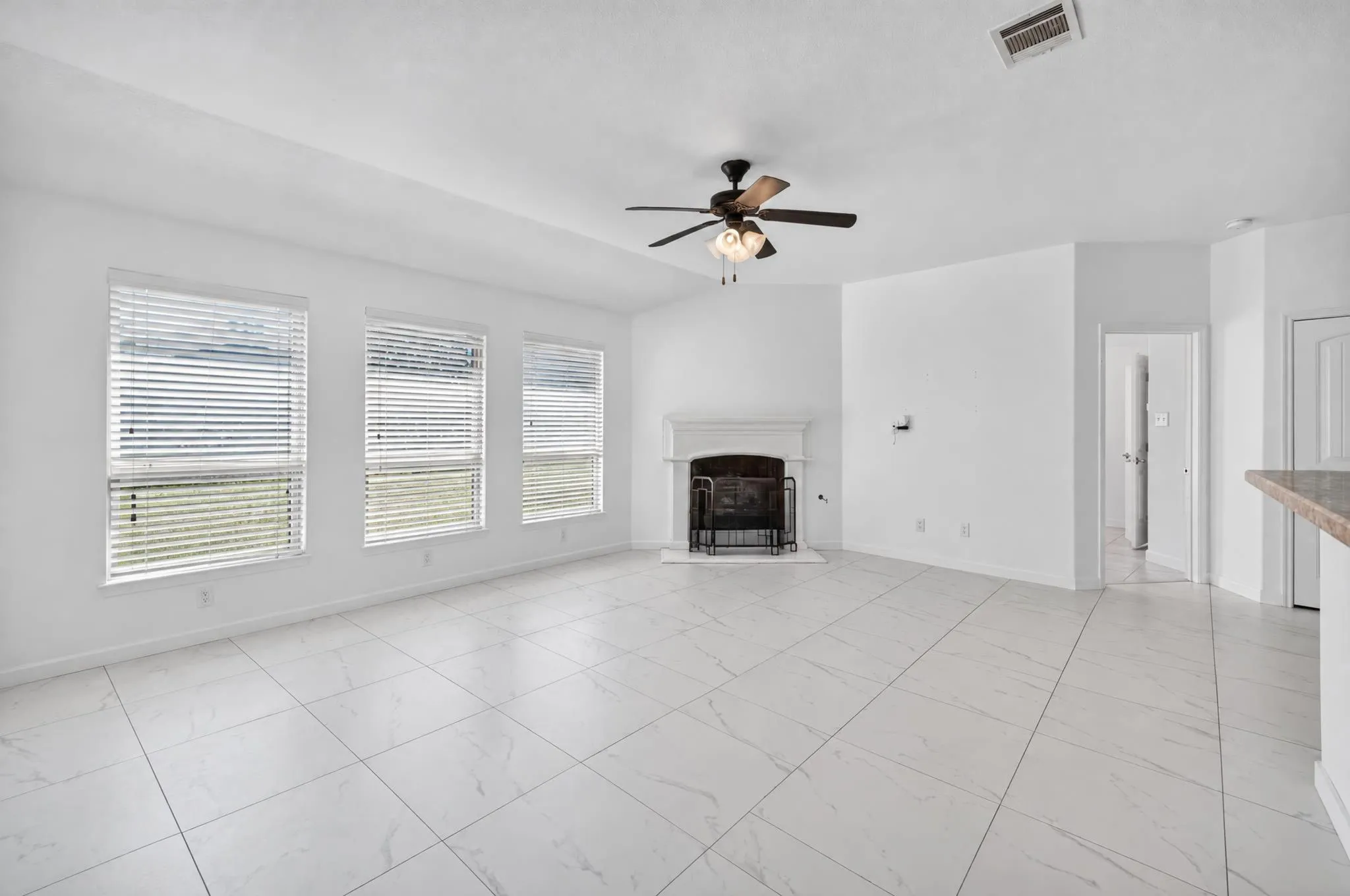 Spacious living area featuring white tile flooring, a fireplace with a white mantel, three large windows, a ceiling fan, and vaulted ceilings