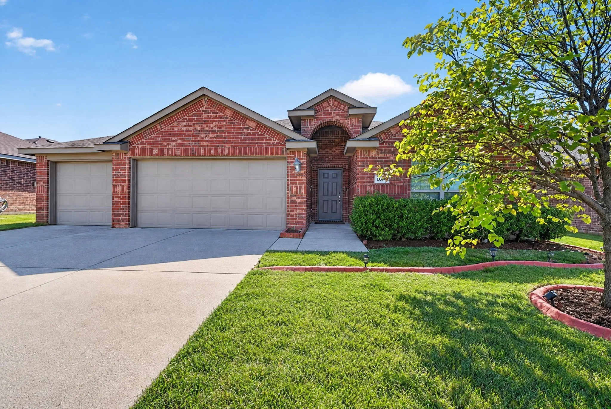 Brick exterior with archway entry, multi-car garage, concrete driveway, and landscaped front yard with mature tree