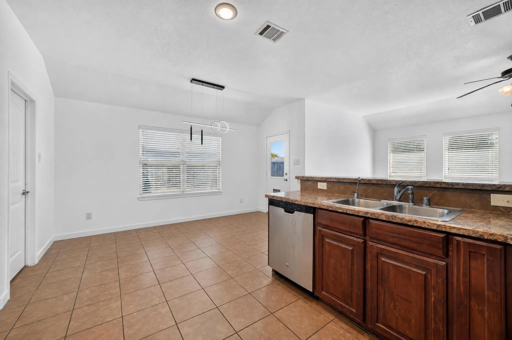 Open-concept living space with tile flooring throughout, featuring a kitchen island with a double basin sink and stainless steel dishwasher, and a dining area with a modern pendant light fixture