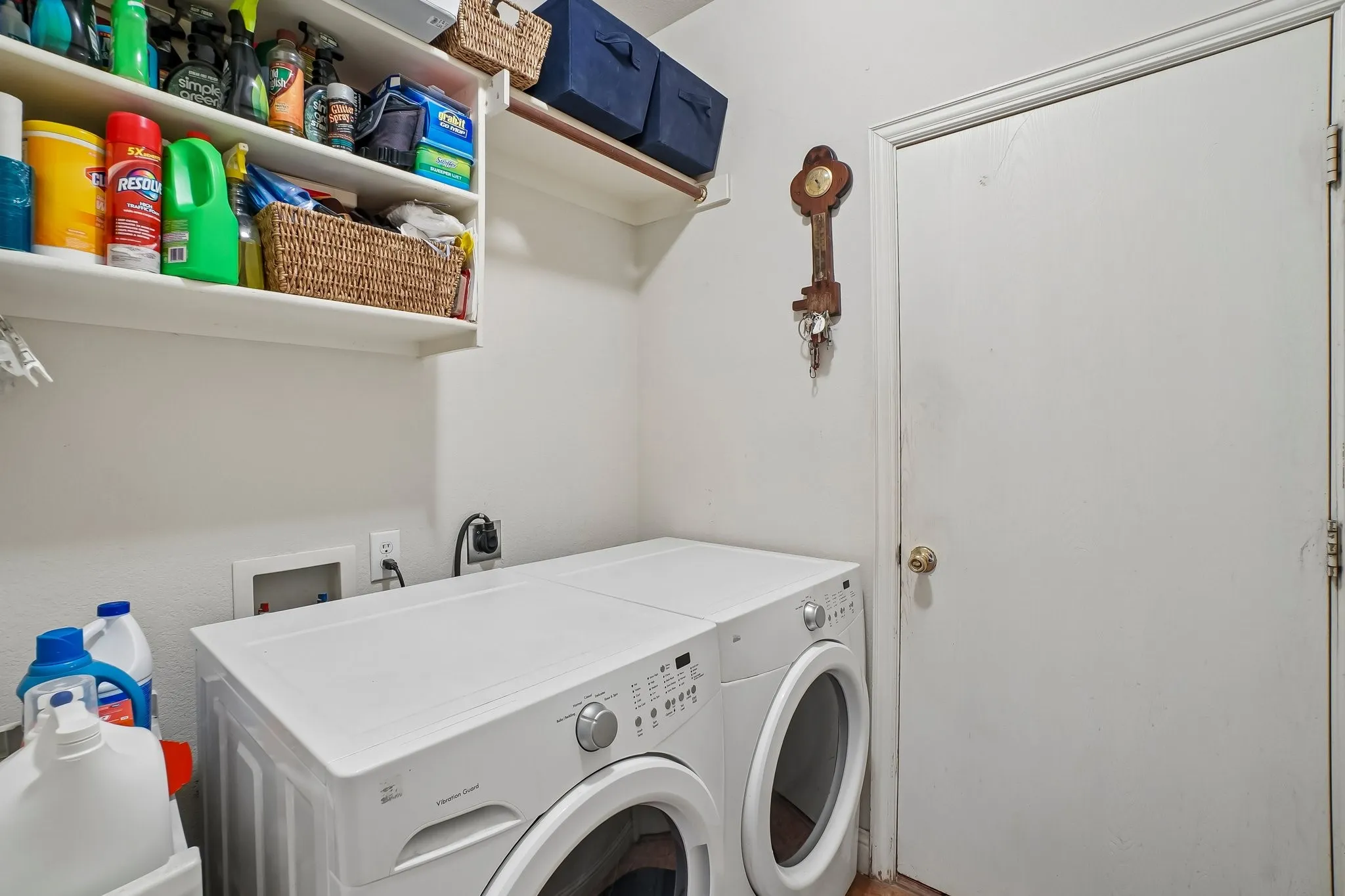 Dedicated laundry room featuring side-by-side front-load washer and dryer with white finish, ample storage shelving with woven baskets and organized supplies, white cabinetry, and direct access to the home's utility areas