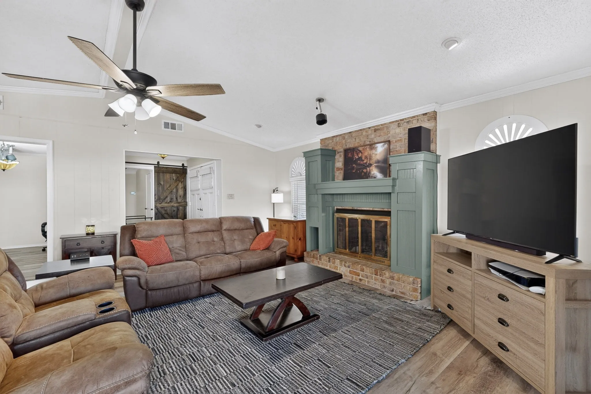 Vaulted ceiling with a ceiling fan, wood-finish flooring, brick-faced fireplace with a painted mantel, decorative window accent, and a barn door entry