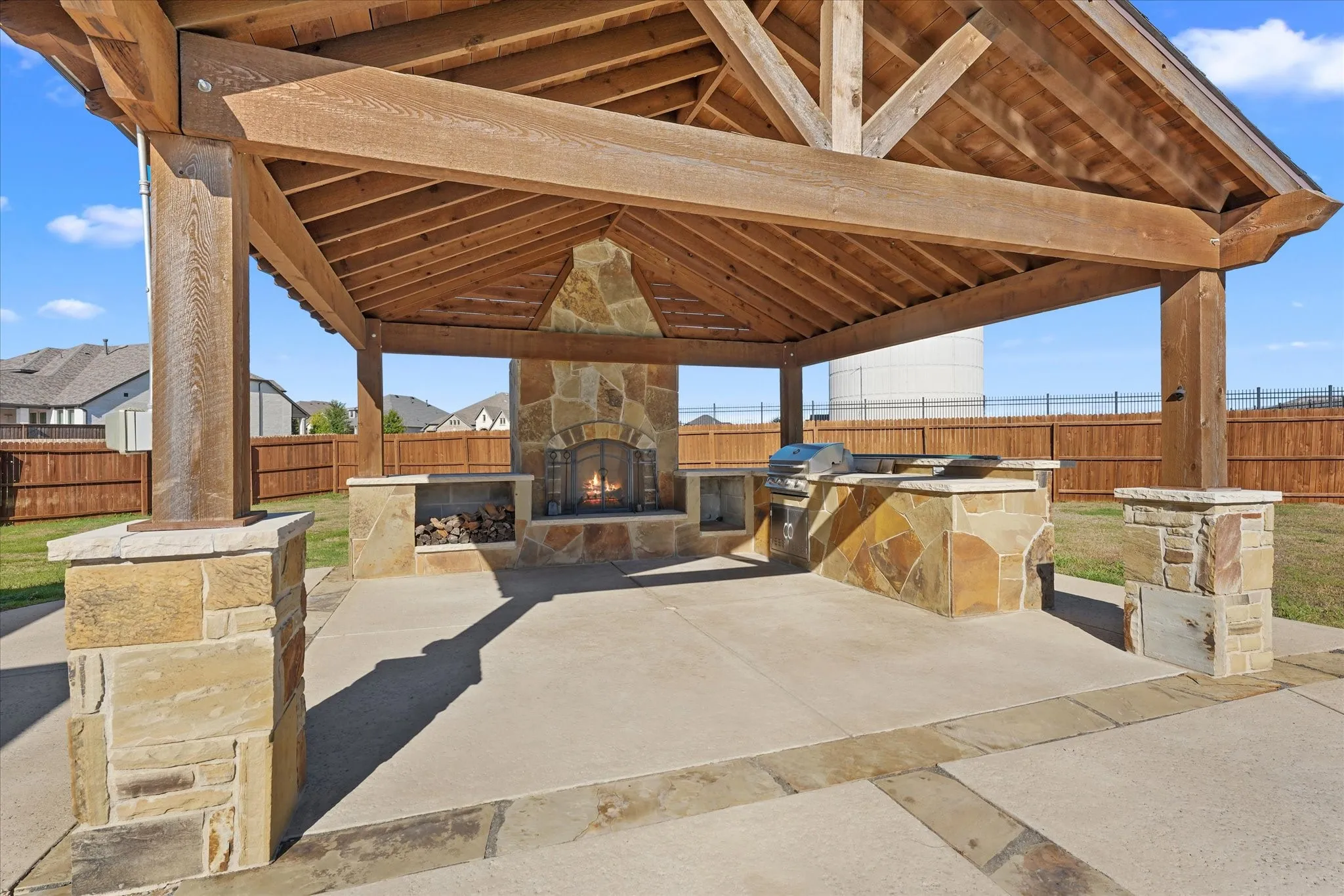 Covered outdoor entertaining area featuring a stone fireplace, built-in grill, stone-clad counter space, and a wood-beamed ceiling