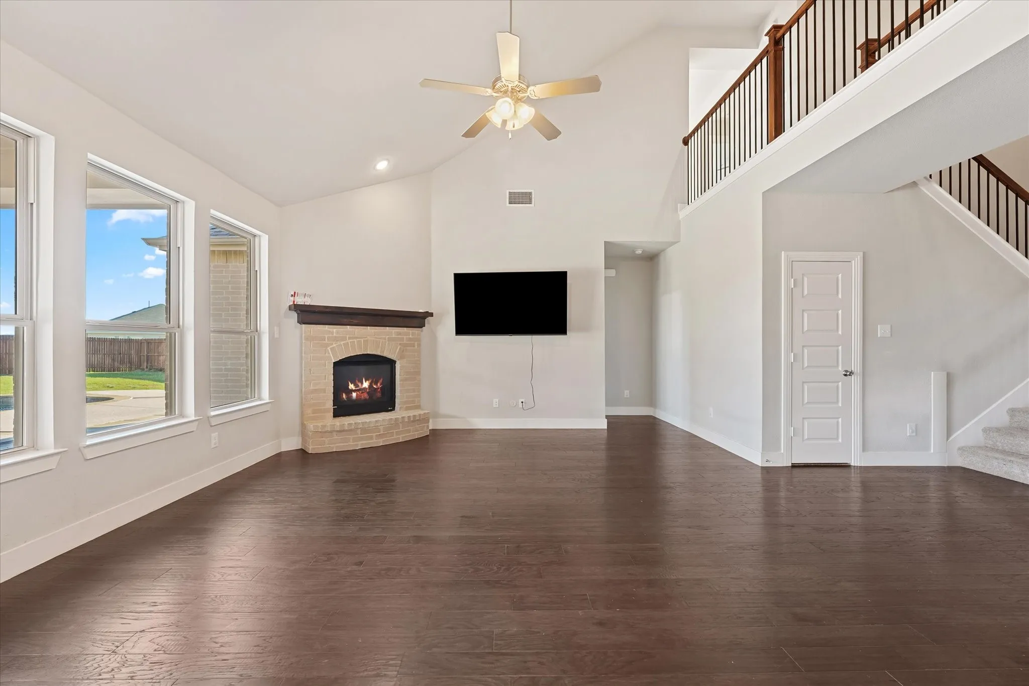 Vaulted ceiling with a ceiling fan, corner fireplace with a brick surround, multiple windows, wood-finish flooring, and a second-story interior balcony with wood and metal railing