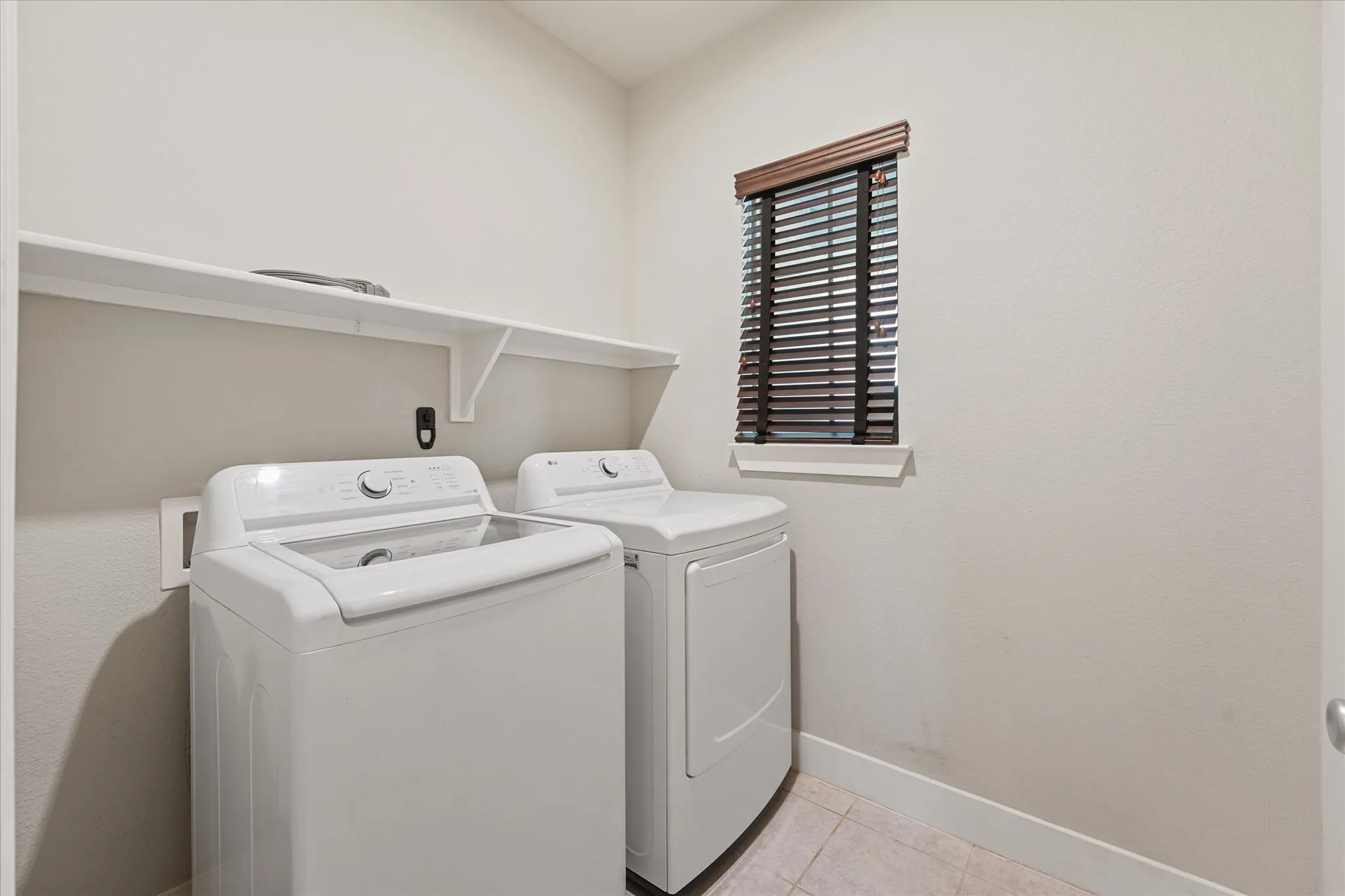 Functional laundry area featuring a window with dark wood-finish blinds, a built-in white shelf, and tiled flooring
