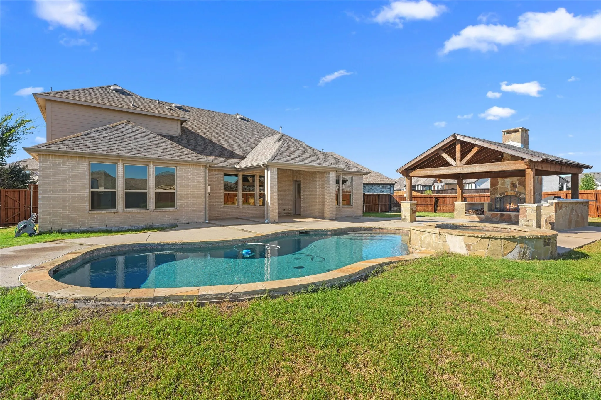 Rear exterior featuring a freeform swimming pool, stone-clad spa, and a covered outdoor kitchen with a stone fireplace