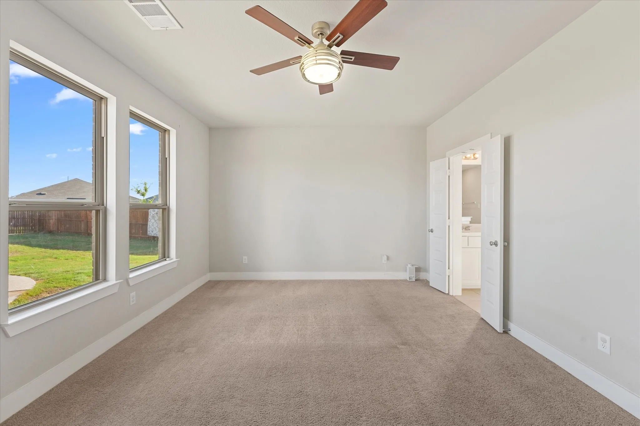 Spacious room featuring neutral carpet flooring, a ceiling fan with integrated lighting, two large windows, and a double-door entry to an additional space with a vanity