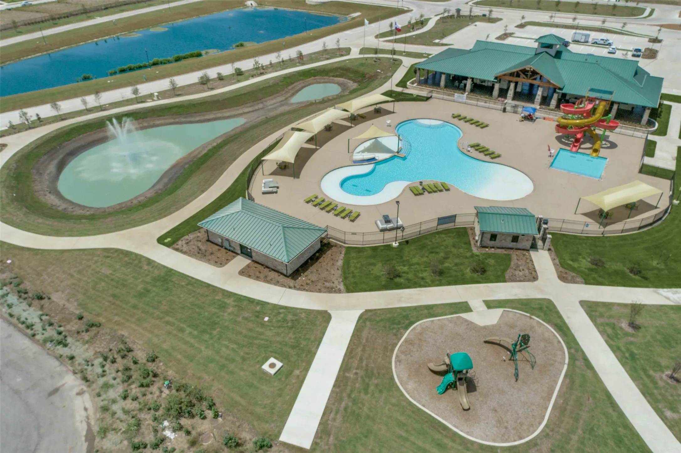 Expansive community amenity center featuring a large swimming pool, separate children's pool with waterslides, a clubhouse with a green roof, a playground, and a decorative pond with a fountain