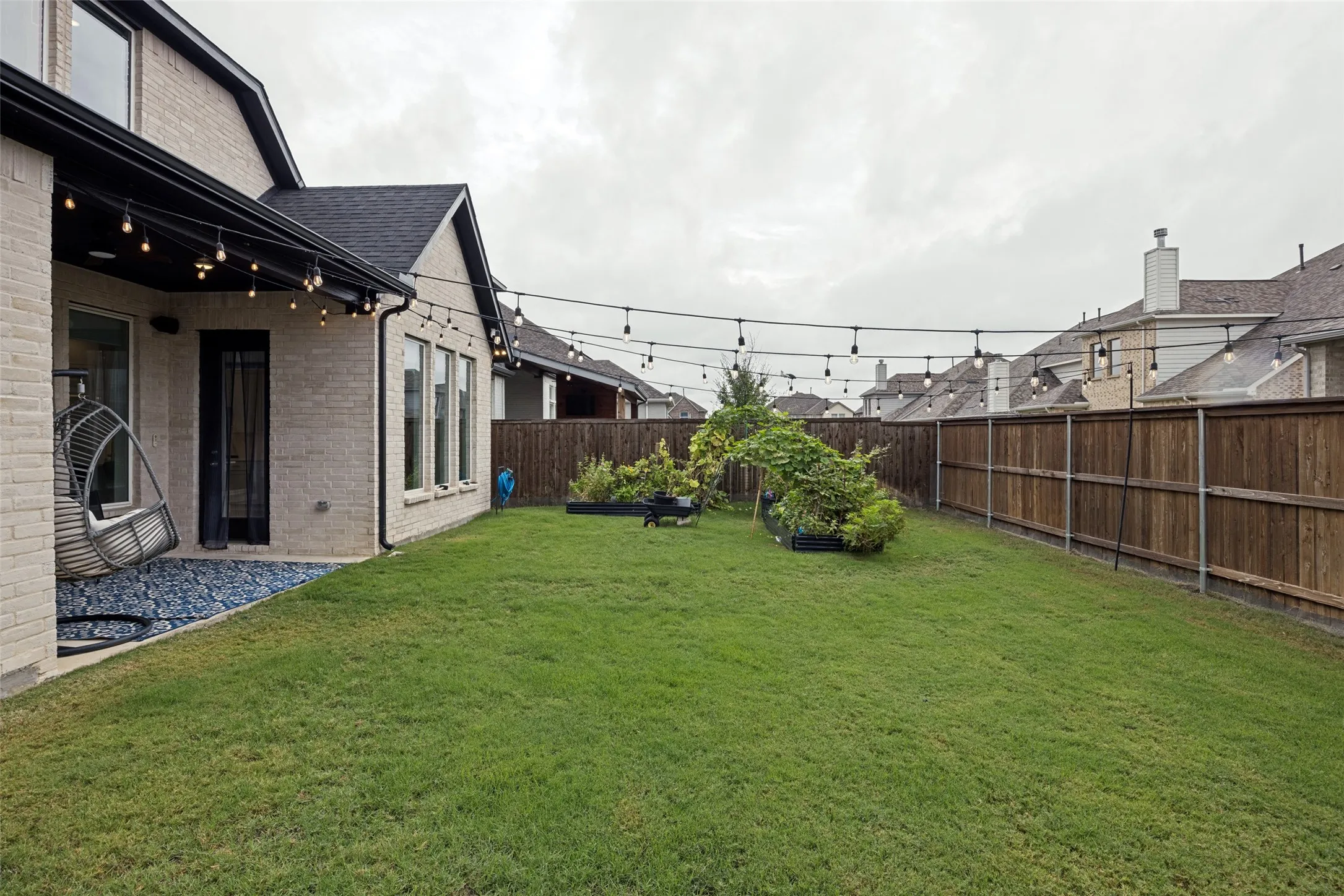 Expansive lawn area with a perimeter wood fence, string lighting, and a stone chip landscaping bed