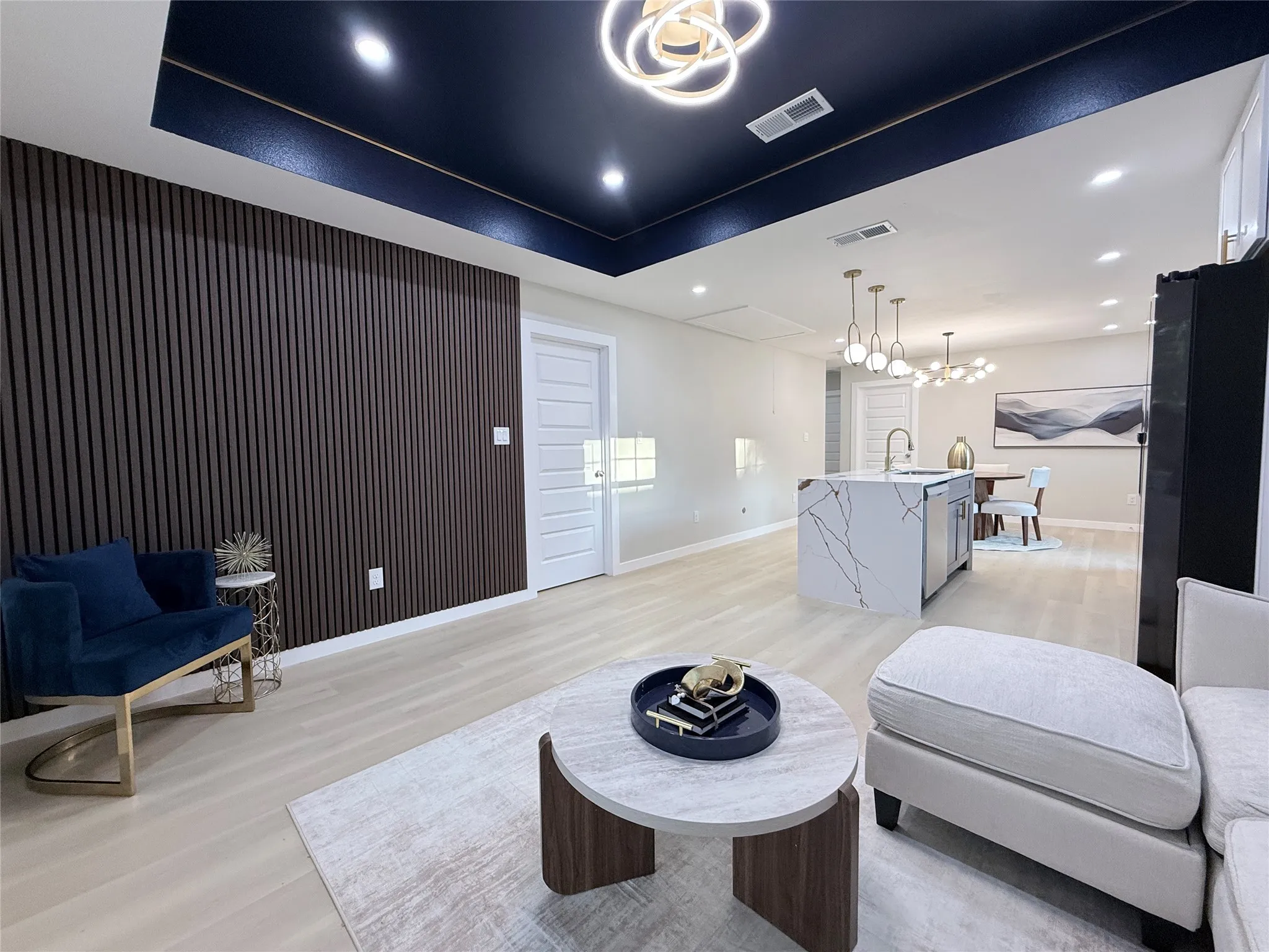 Dark wood-slat accent wall, recessed ceiling with perimeter lighting, light wood-finish flooring, kitchen island with waterfall counter, and modern pendant lighting