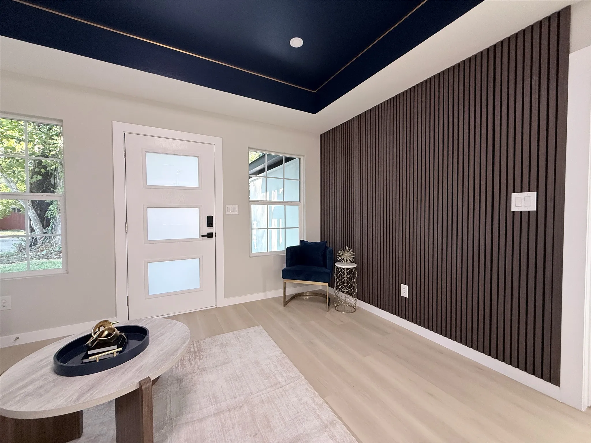 Dark wood panel accent wall, light wood-finish flooring, white entry door with frosted glass panels, and a dark blue ceiling with gold trim