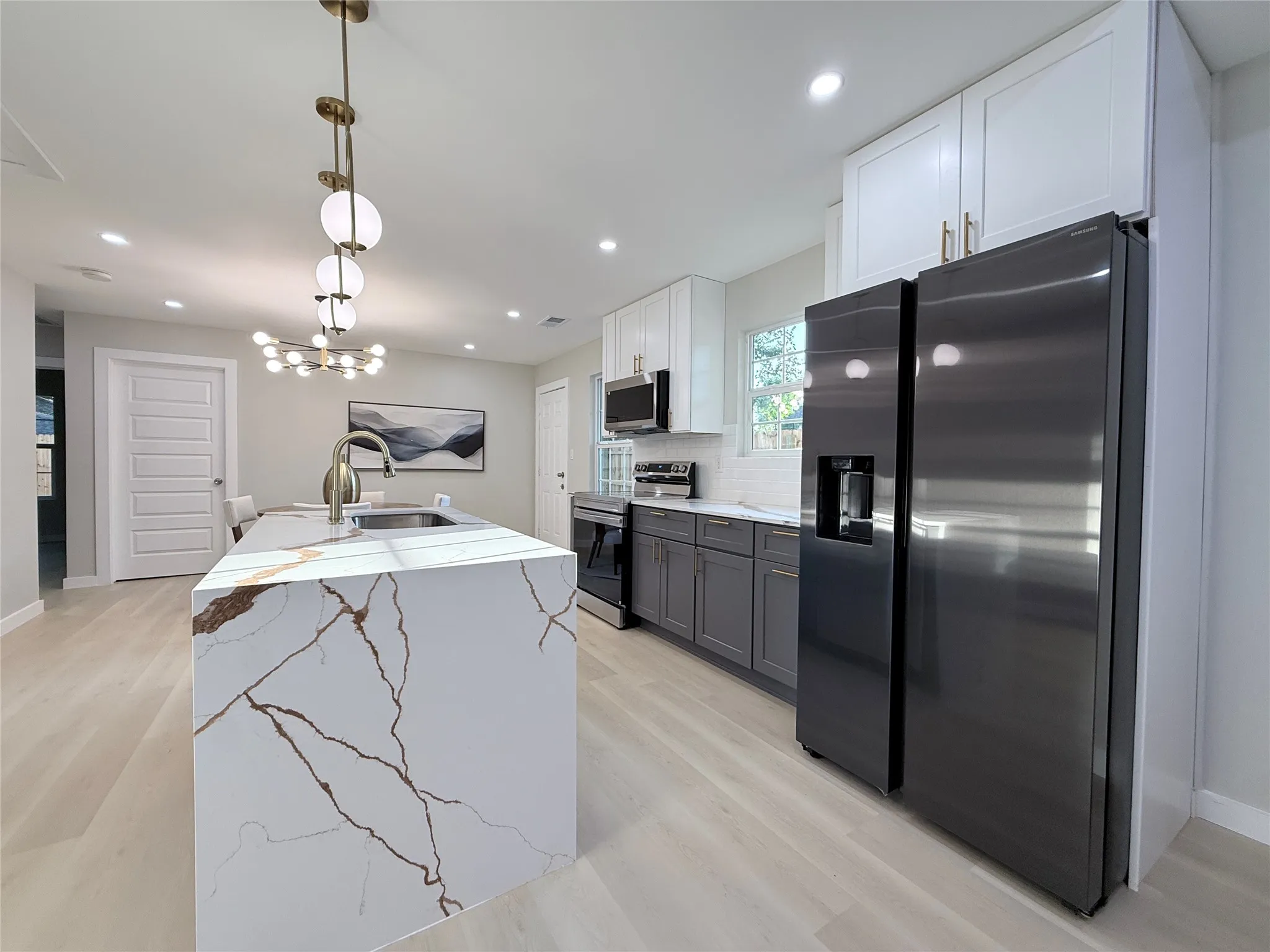 Kitchen featuring a large waterfall island with integrated sink, stainless steel appliances, two-tone cabinetry, wood-finish flooring, and recessed lighting