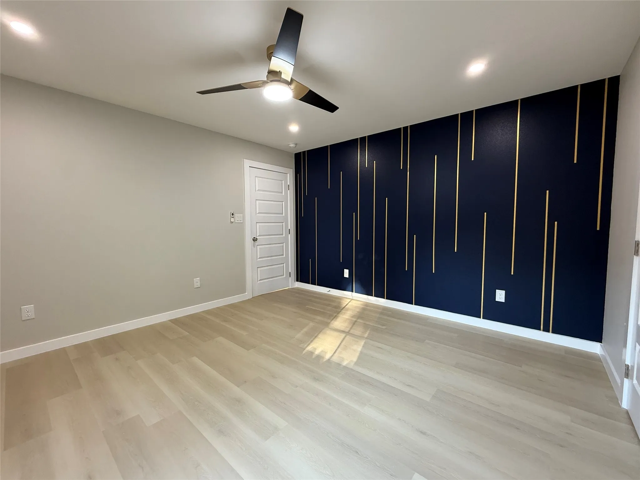 Modern ceiling fan with integrated lighting, recessed ceiling lights, light wood-finish flooring, a paneled interior door, and a navy accent wall with vertical gold detailing
