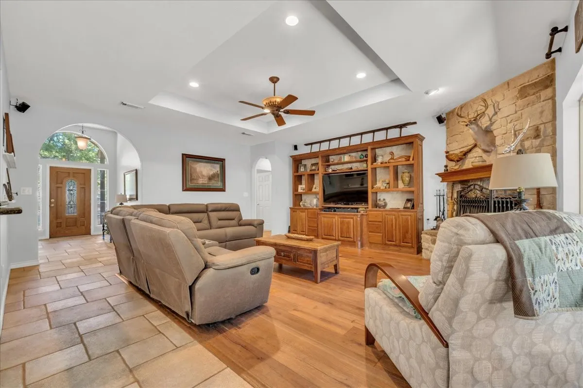 Tray ceiling with recessed lighting and a ceiling fan, stone-clad fireplace, wood-finish flooring, built-in entertainment center with shelving and cabinetry, and a wood-paneled front door with arched transom