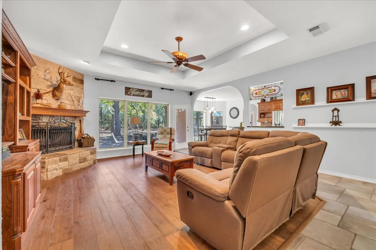 Stone-clad fireplace with a wood mantle, wood-finish flooring, a tray ceiling, and a ceiling fan