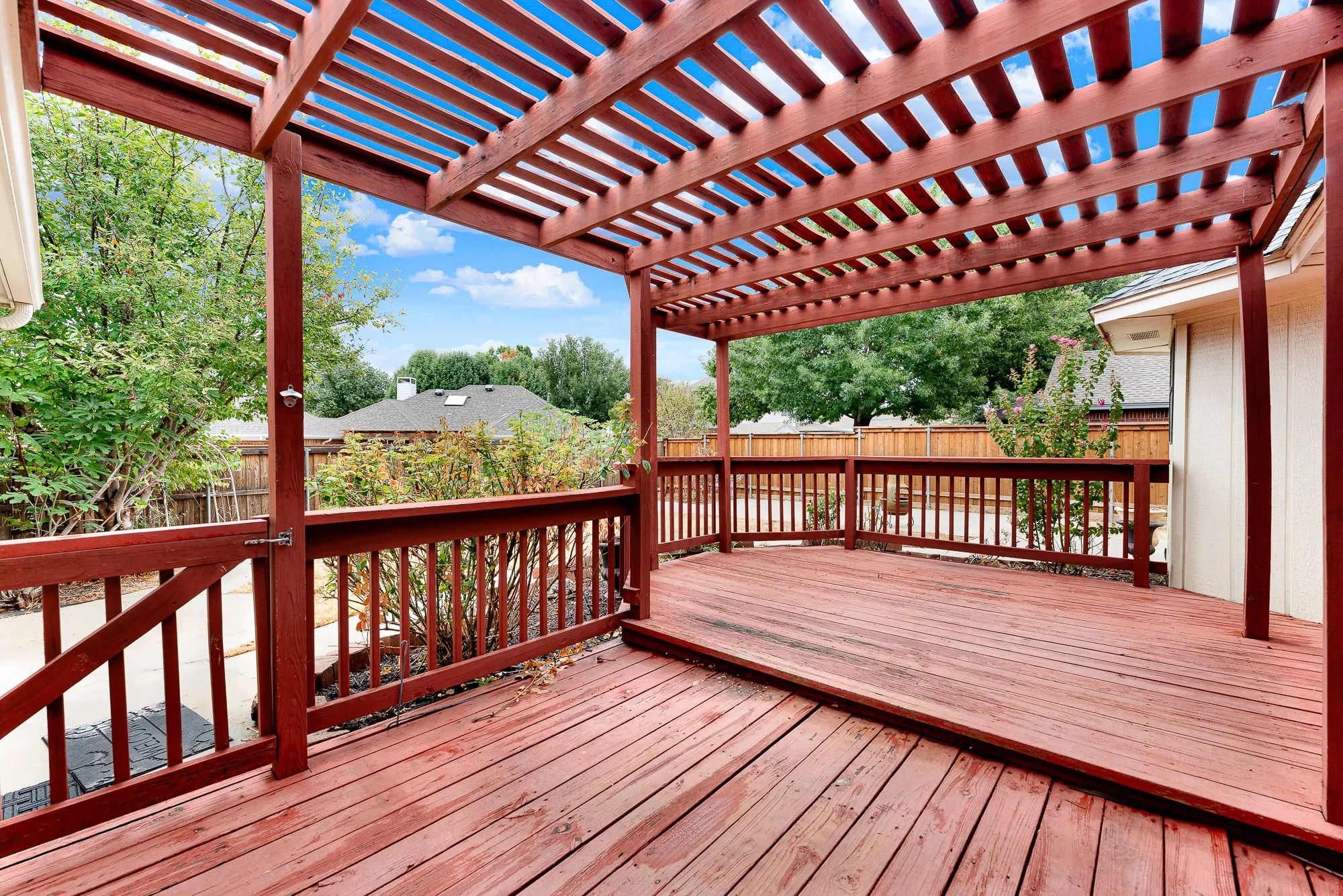 A rich wooden pergola weaves overhead, casting slatted shadows across the multi-tiered deck space. Picture sipping iced tea on a sunny afternoon while a gentle breeze rustles through the garden beds.