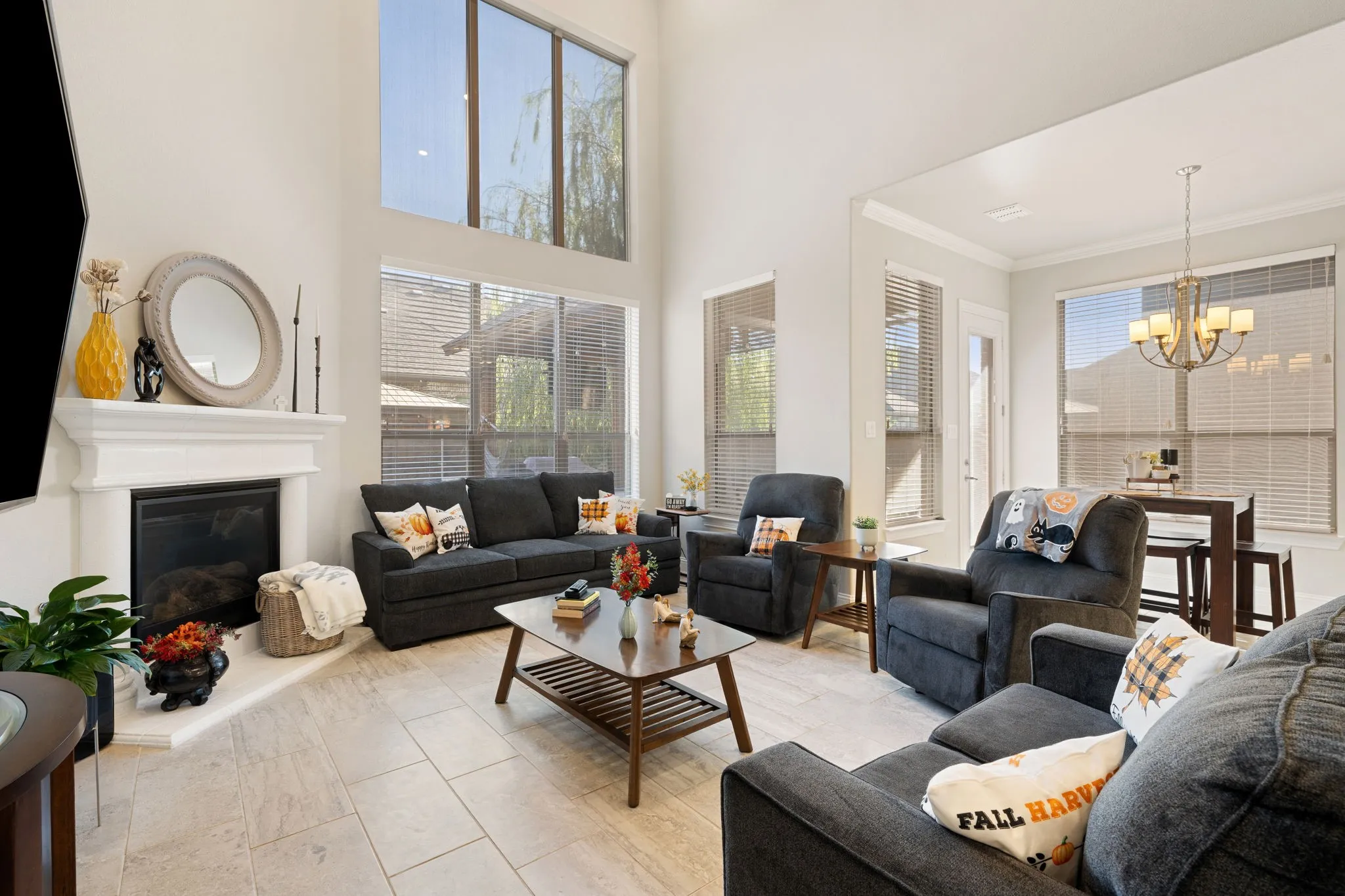 Vaulted ceilings with high windows, a fireplace with a white mantel, light-toned tile flooring, and multiple windows with blinds