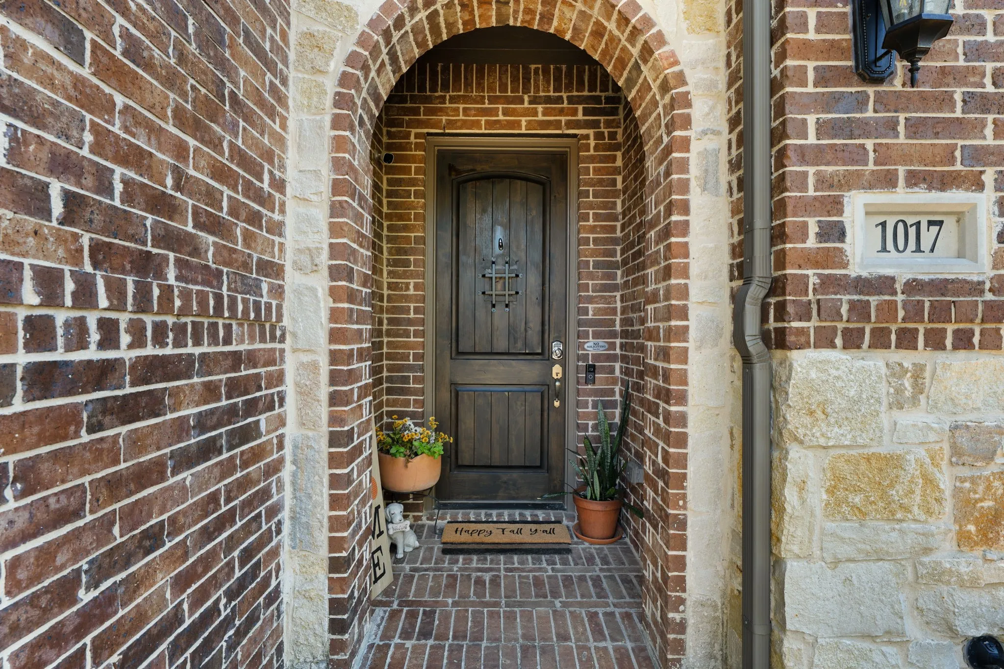 Arched brick entryway featuring a dark wood-finish door with decorative ironwork, brick and stone exterior, and a stone house number plaque