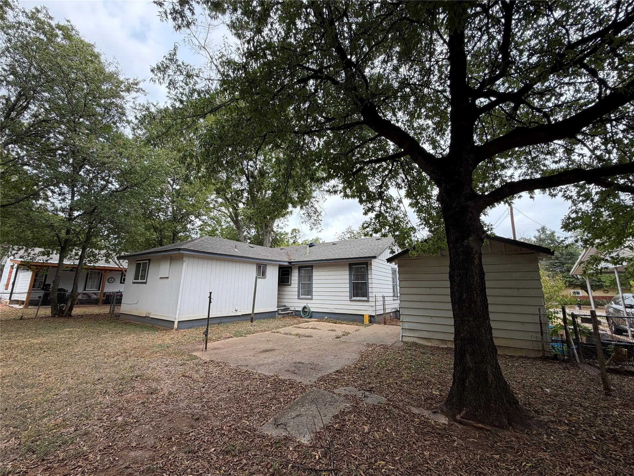 Rear exterior featuring white horizontal siding, a gray shingle roof, a concrete patio, and mature trees