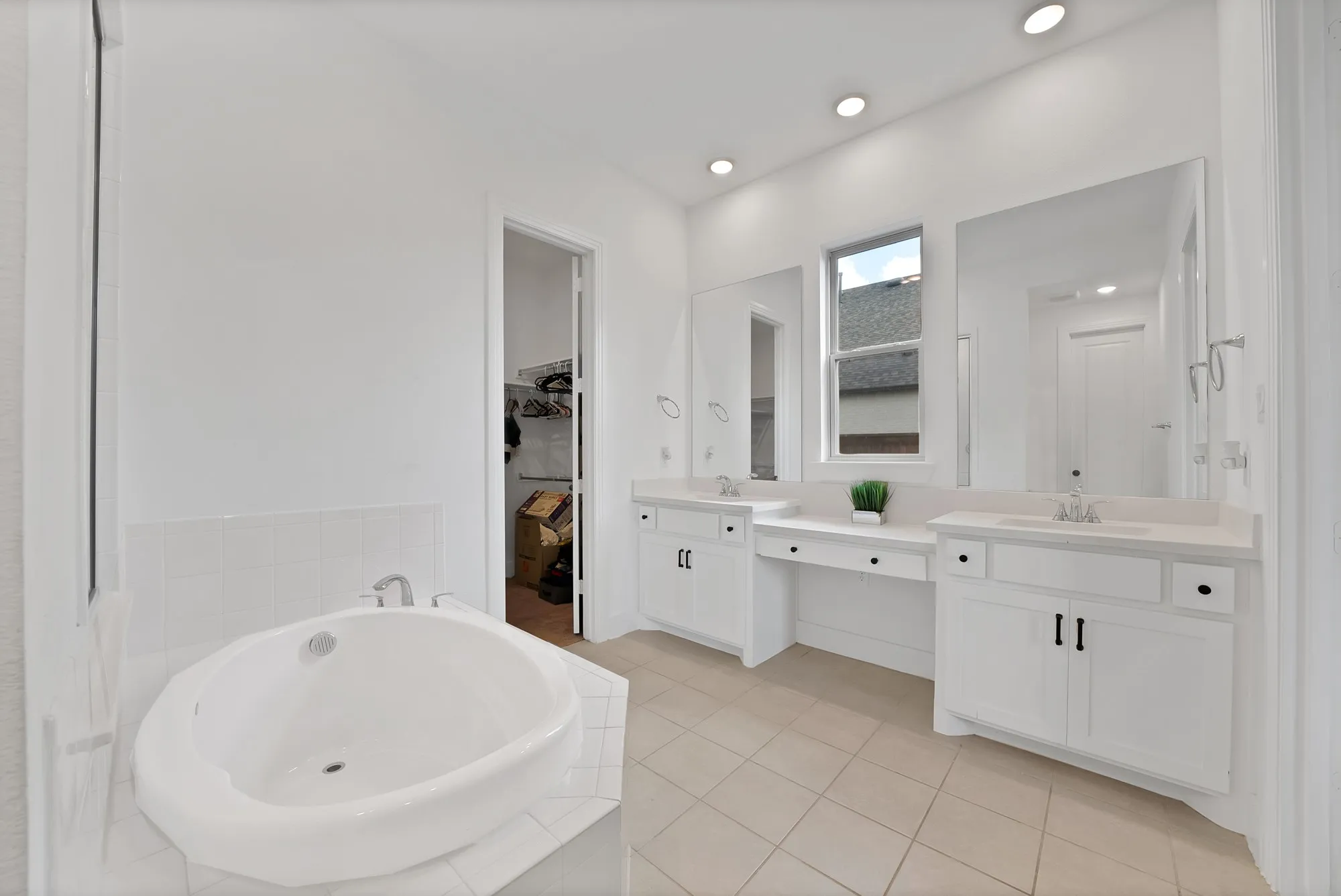 Built-in soaking tub, dual vanity with white cabinetry, light-toned tile flooring, recessed lighting, and a window for natural illumination