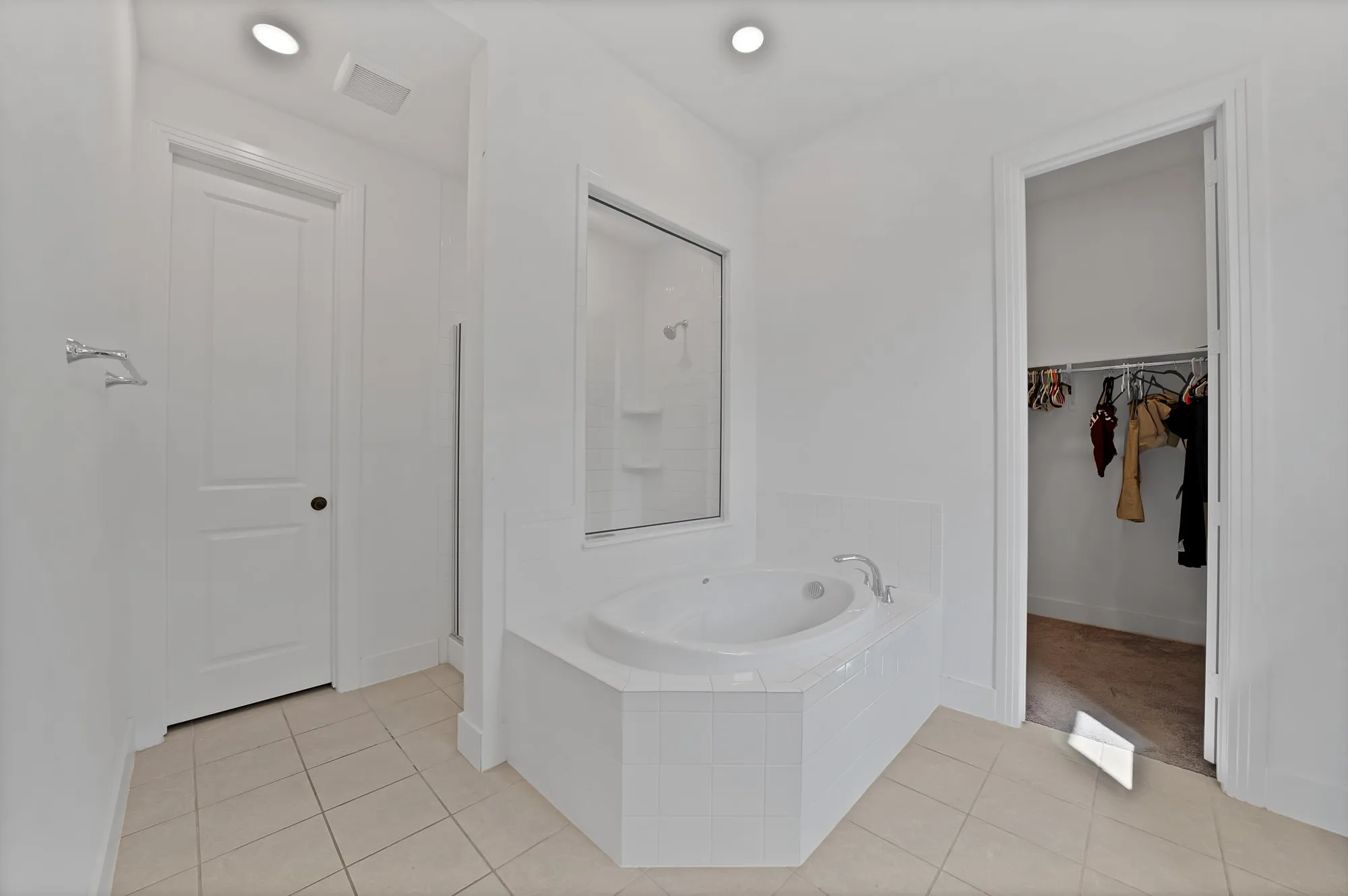 Recessed lighting illuminates a corner jetted tub, a separate shower with a clear glass enclosure, and a walk-in closet with hanging rails