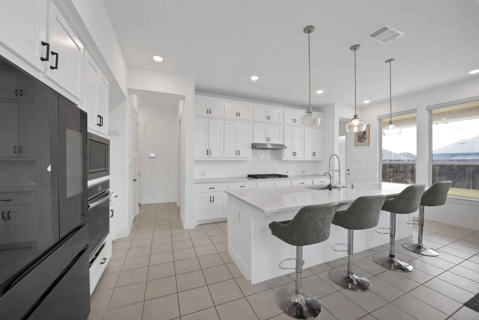 White cabinetry with black hardware, tile flooring, kitchen island with built-in sink, stainless steel range hood, and multiple windows providing natural light