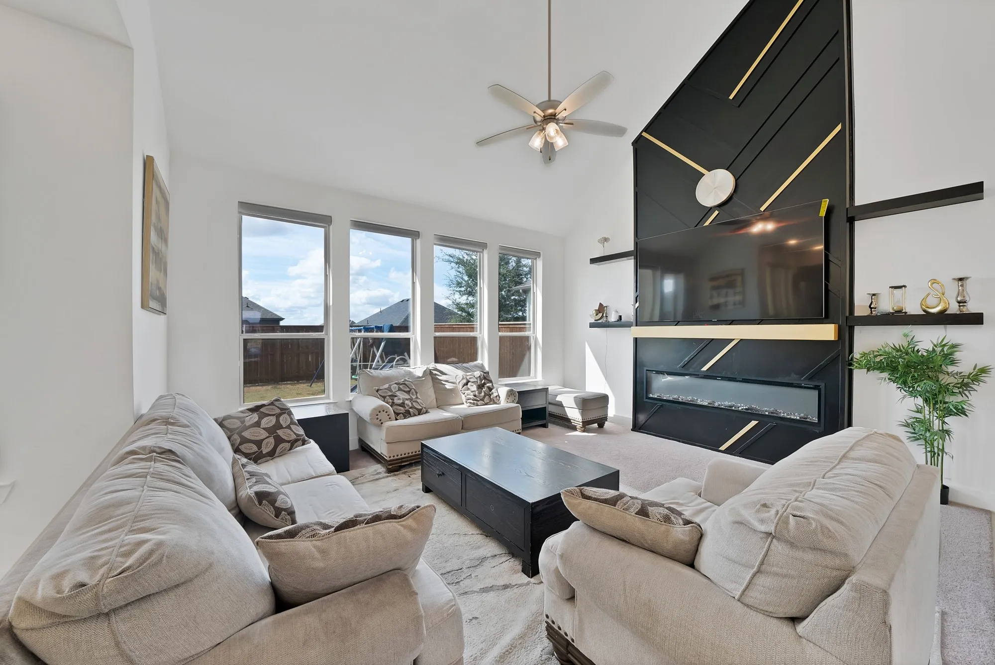 Vaulted ceiling with a ceiling fan, three large vertical windows, a built-in electric fireplace, and decorative wall shelving