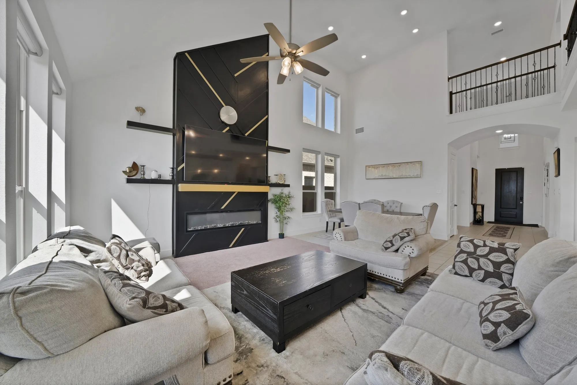 Vaulted ceilings with recessed lighting, black fireplace wall with gold accents, floor-to-ceiling windows, second-story interior balcony with wrought iron railing, and a ceiling fan with light fixture