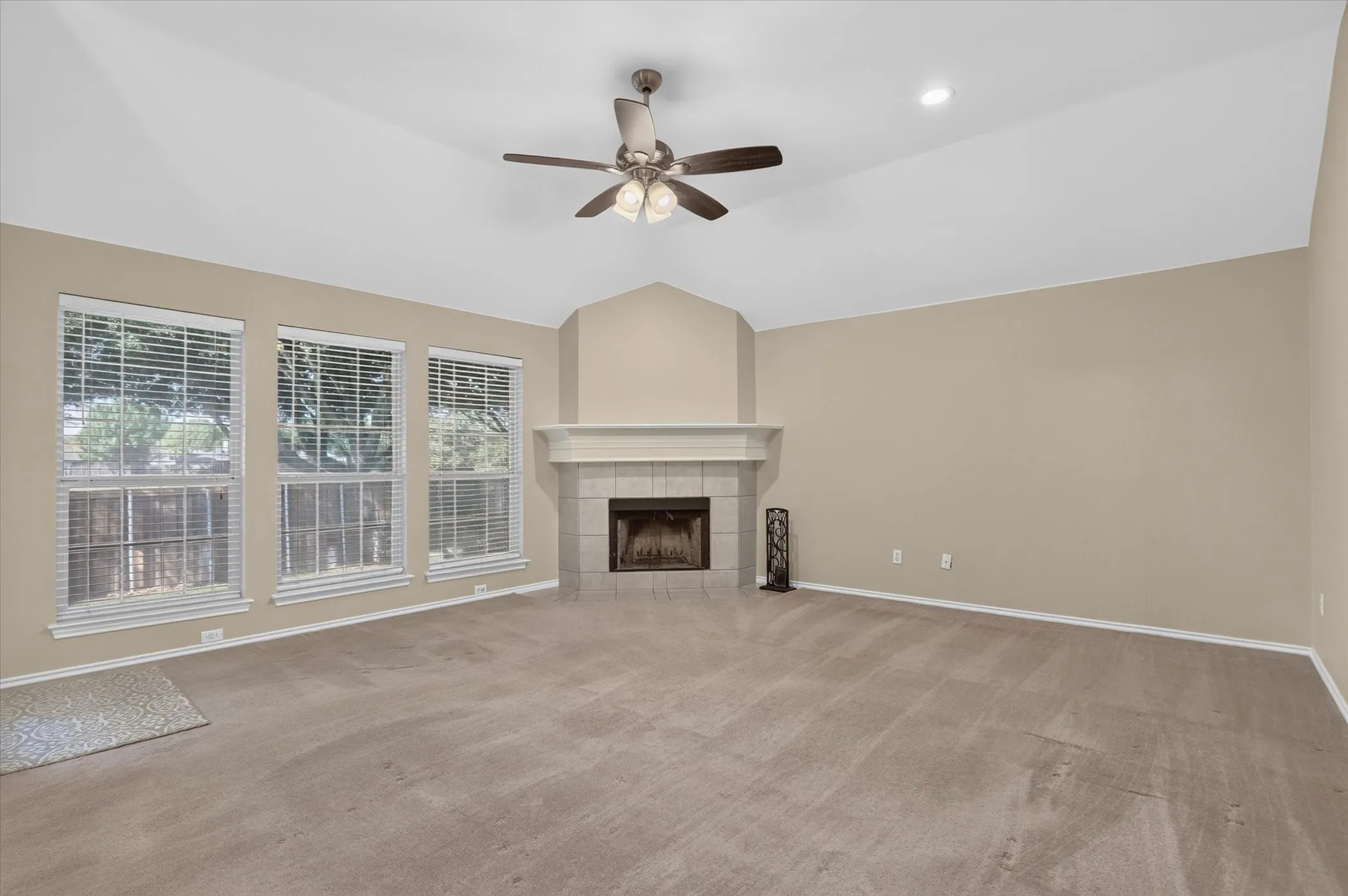 Vaulted ceiling with a ceiling fan, three large windows, corner fireplace with a mantel, and neutral carpeting
