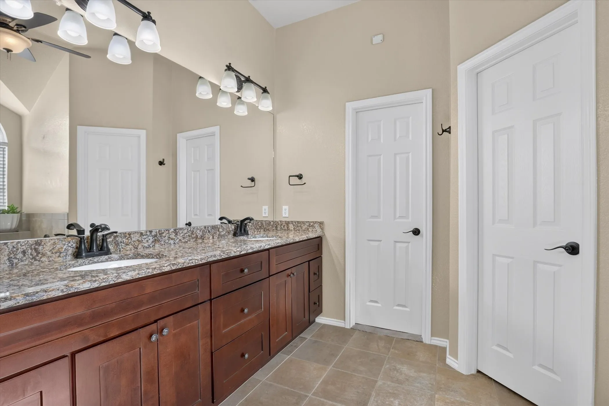 Dual sinks set in a stone-finish countertop, extensive wood-finish cabinetry, full-width mirror, and tiled flooring
