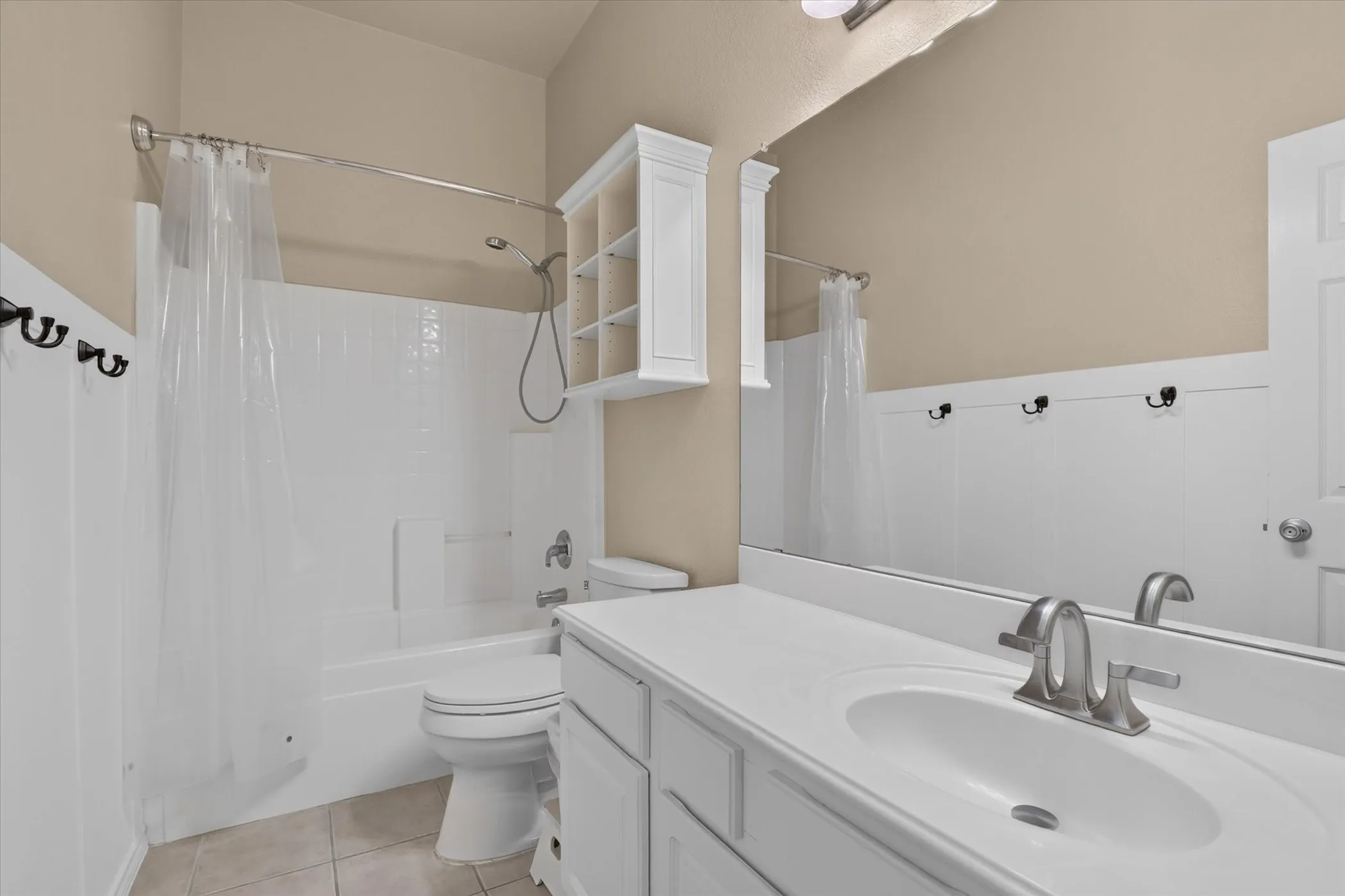 White vanity with an integrated sink and brushed nickel faucet, full-width mirror, white paneled wainscoting, white bathtub and shower combination, and tile flooring