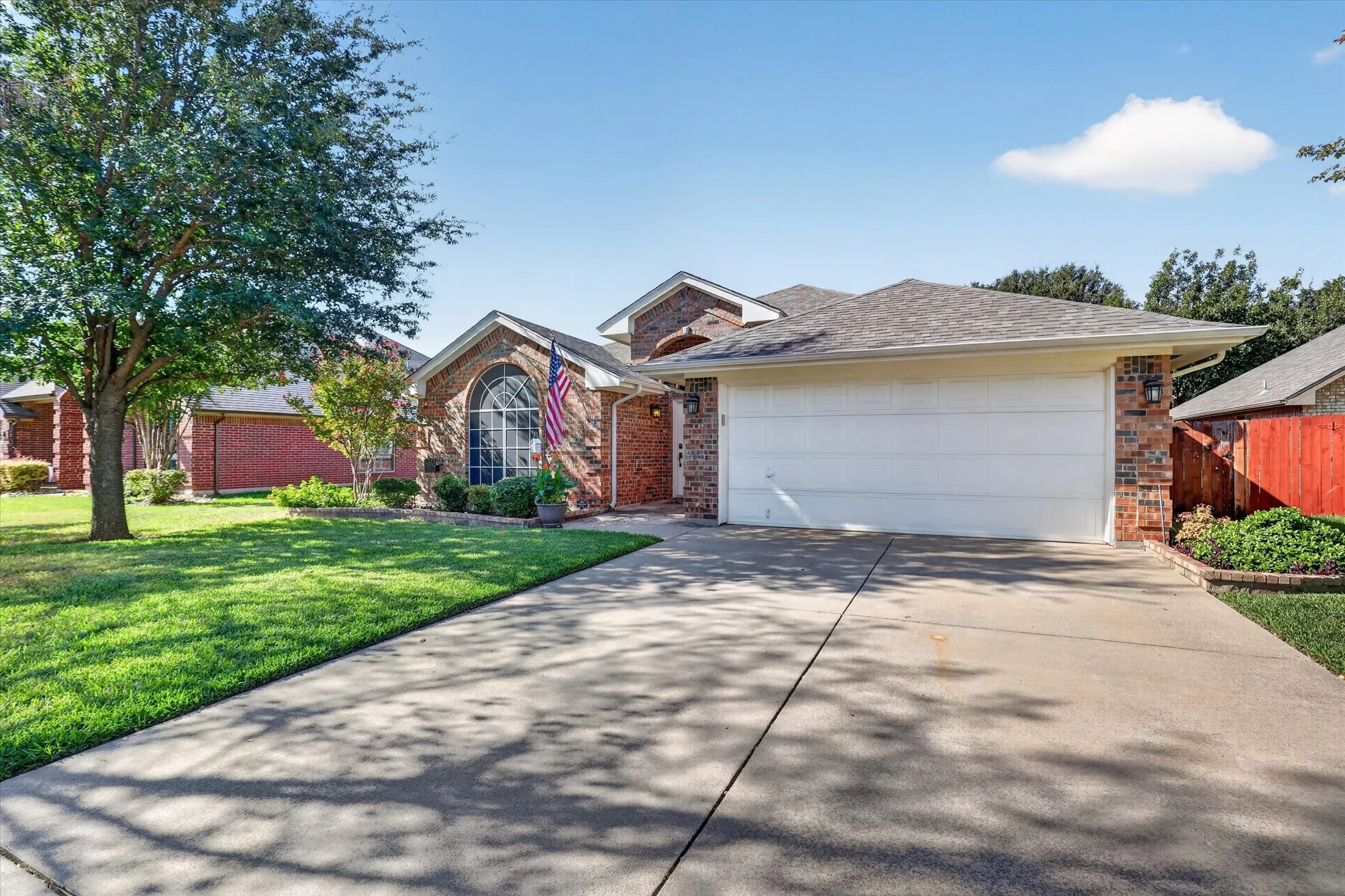 Brick exterior with an arched window, attached garage, concrete driveway, and established landscaping