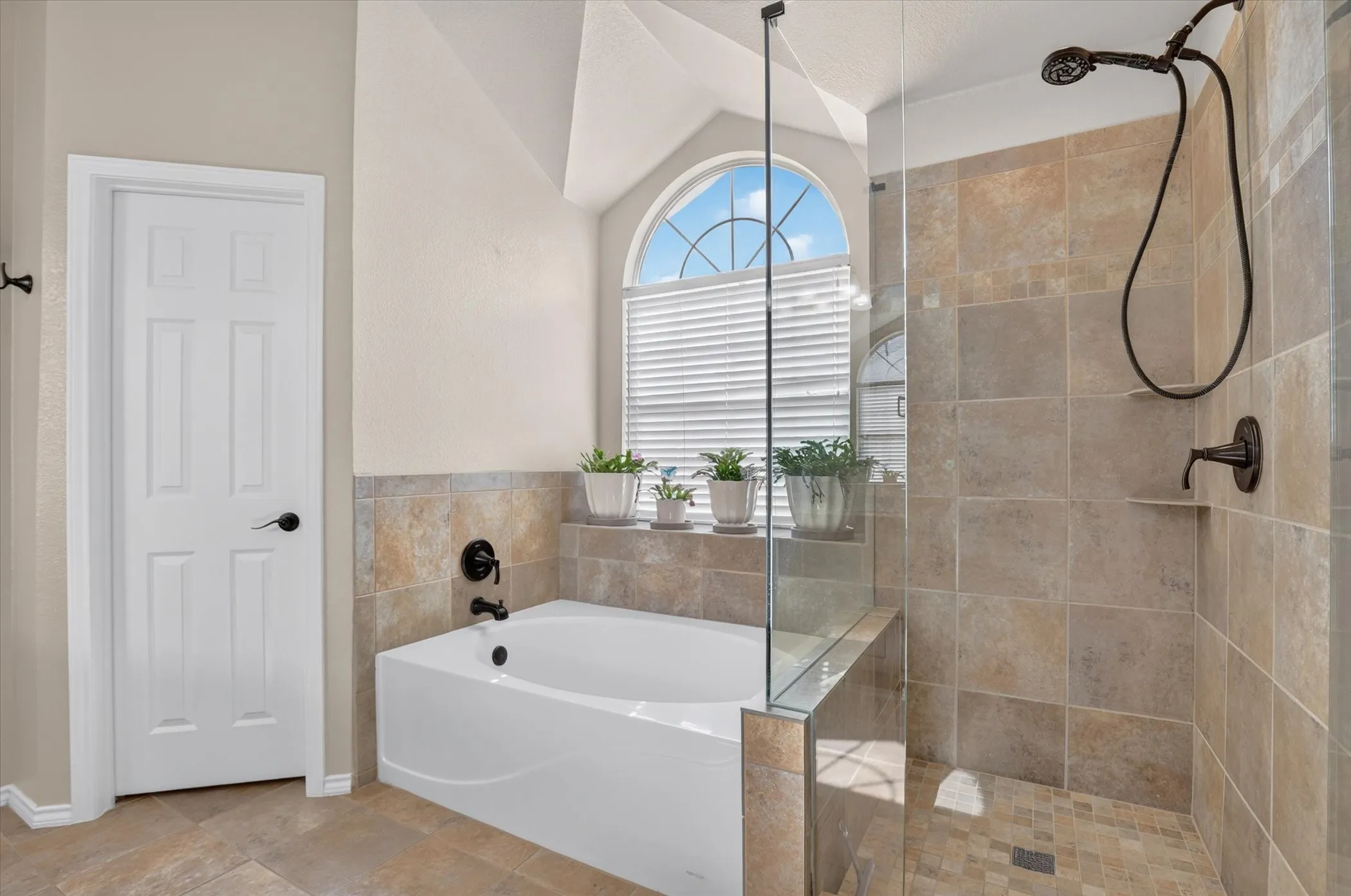 Freestanding soaking tub with dark bronze fixtures, framed by a curved window with privacy blinds