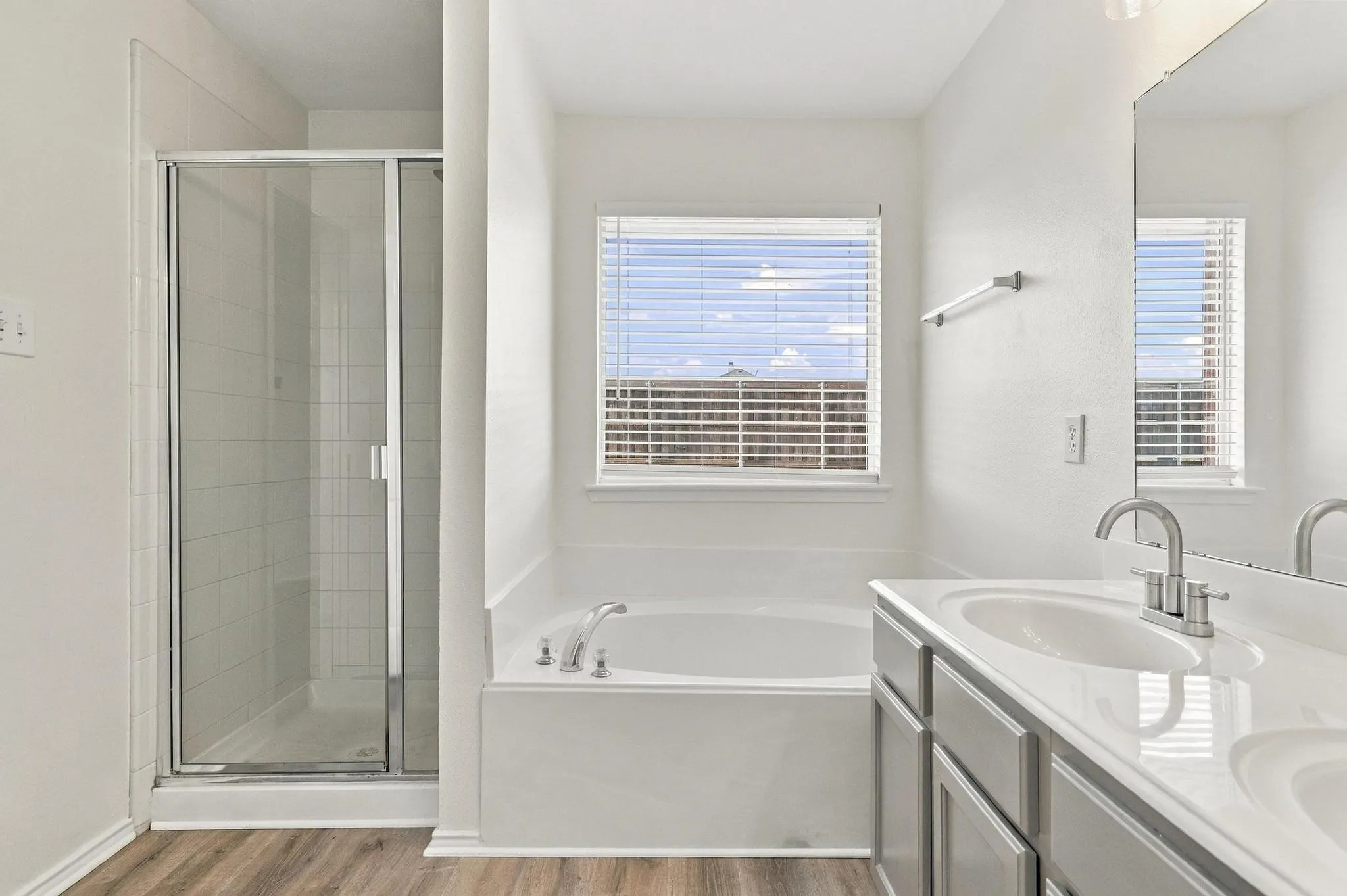 Dual vanity with undermount sinks, wood-finish flooring, a built-in tub, a separate glass-enclosed shower, and a large window