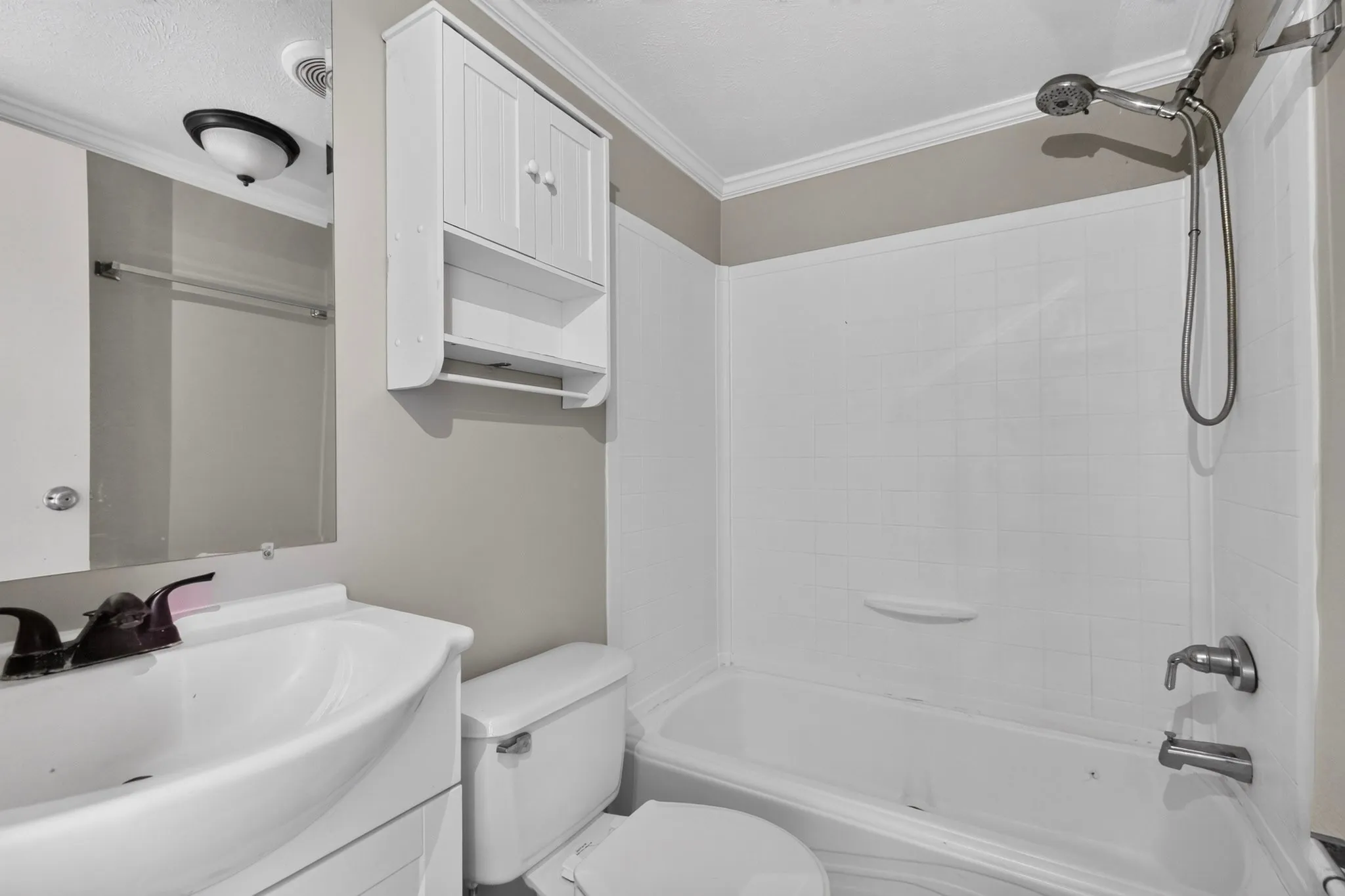 White vanity with integrated sink, wall-mounted mirror, over-toilet storage cabinet, and a shower-over-tub combination with a handheld shower fixture