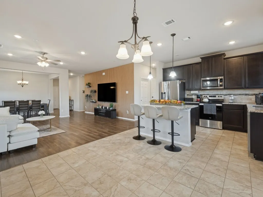 Open-concept living area featuring wood-finish flooring throughout, a centered kitchen island with seating for multiple guests, stainless steel appliances, dark cabinetry with contrasting countertops, pendant lighting, recessed ceiling lights, and a ceiling fan