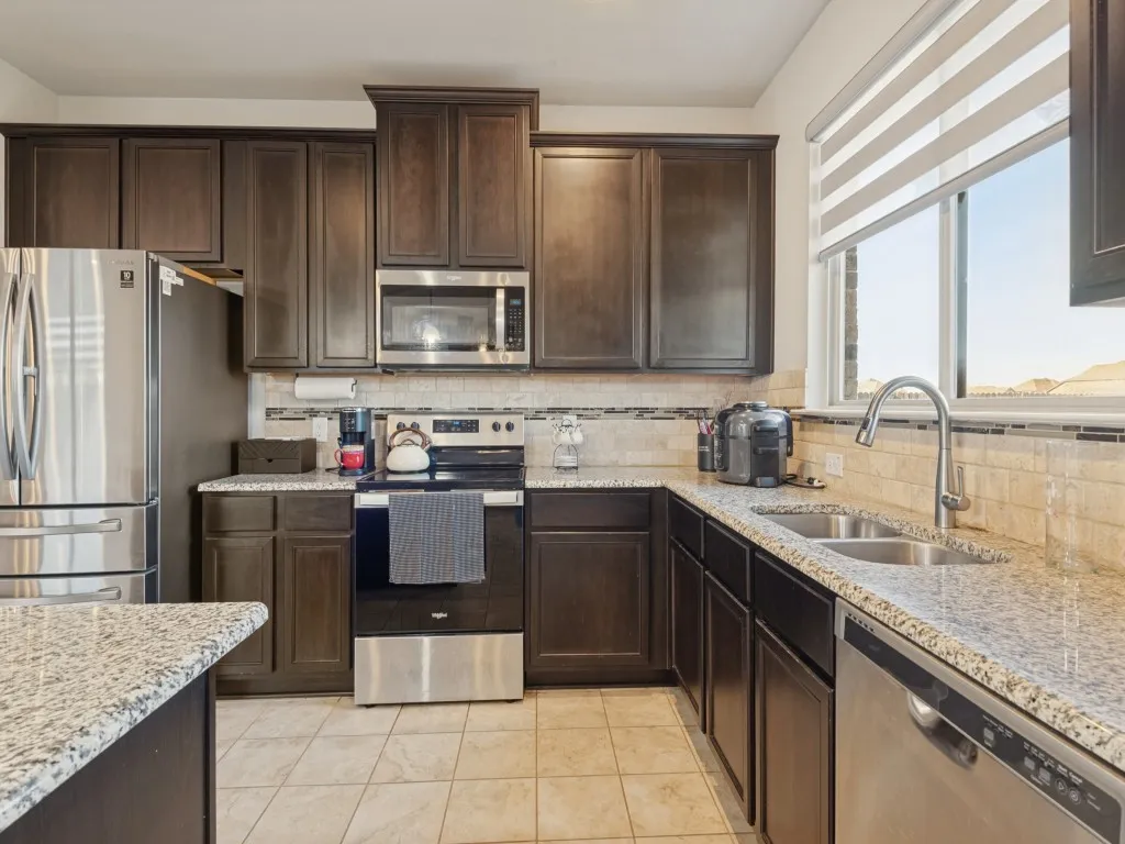 Dark wood-finish cabinetry with crown molding, stainless steel appliances, granite countertops, tile backsplash, and a double basin stainless steel sink