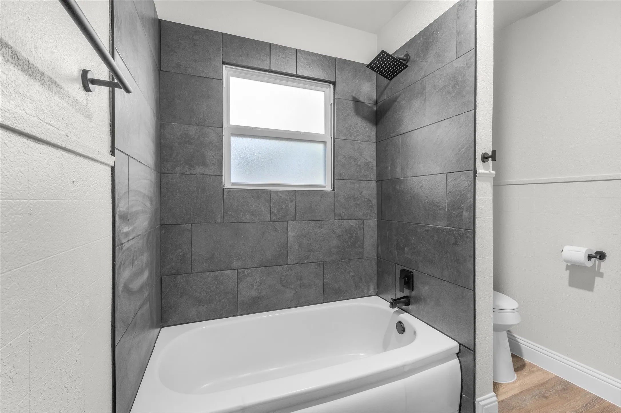 Bathtub with full-height gray tile surround, matte black shower fixtures, and an opaque window