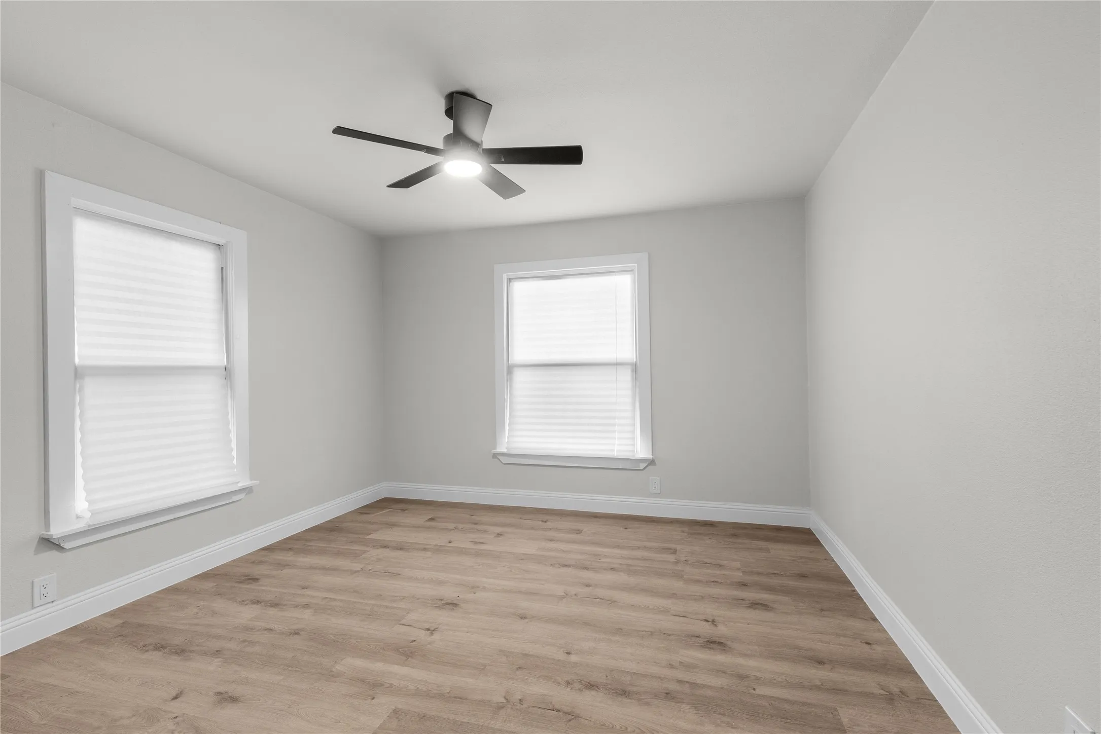 Bright corner bedroom with abundant natural light from two large double-hung windows, wood-finish flooring, and ceiling fan with integrated lighting fixture