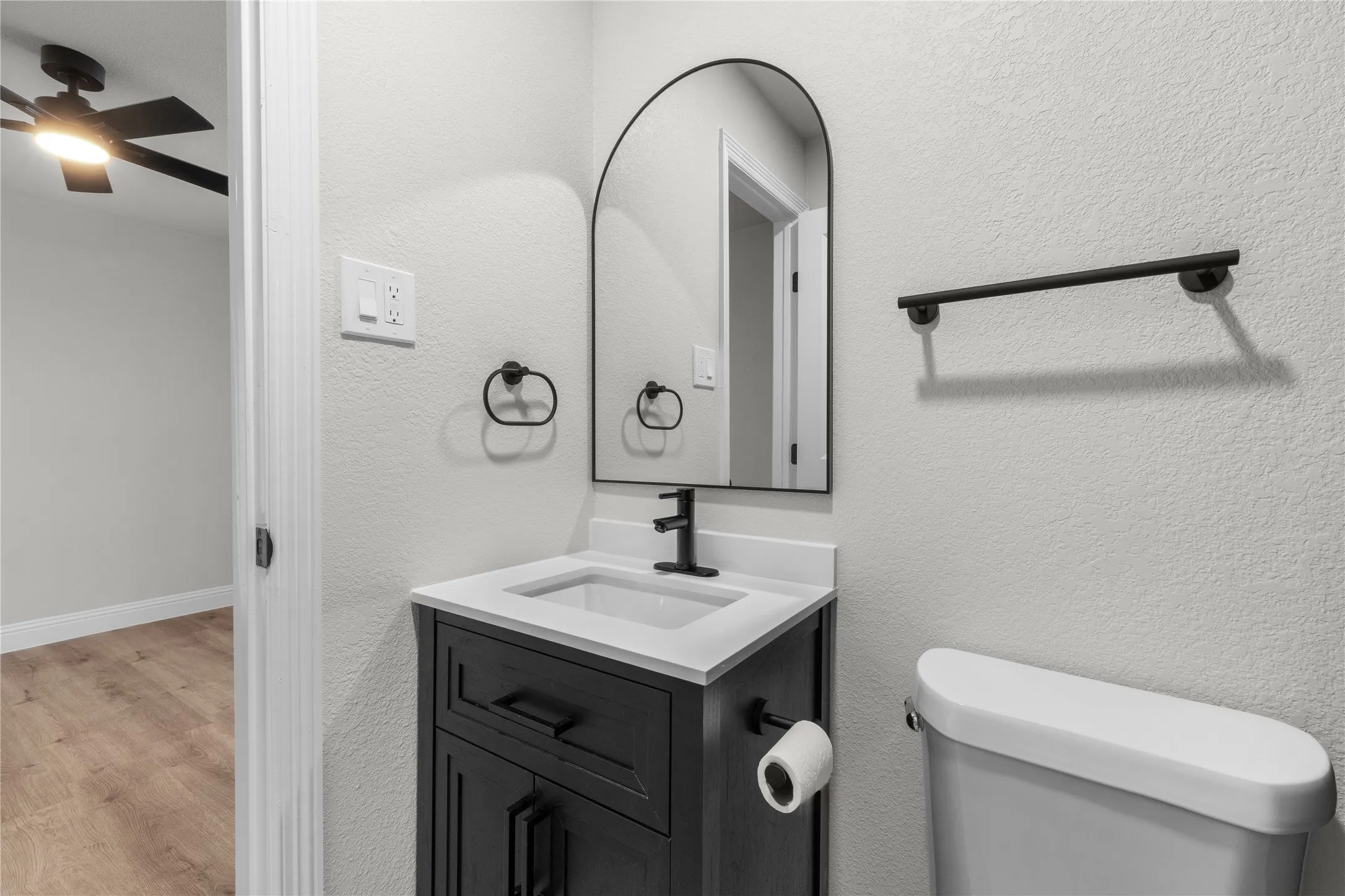Bathroom featuring a dark wood-finish vanity with a white countertop and integrated sink, black hardware, an arched mirror, and a ceiling fan visible in the background