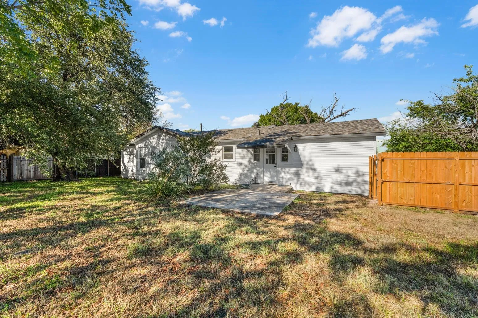 Spacious backyard with mature tree canopy and expansive lawn, complemented by a single-story white exterior home featuring a wooden privacy fence and detached storage structure