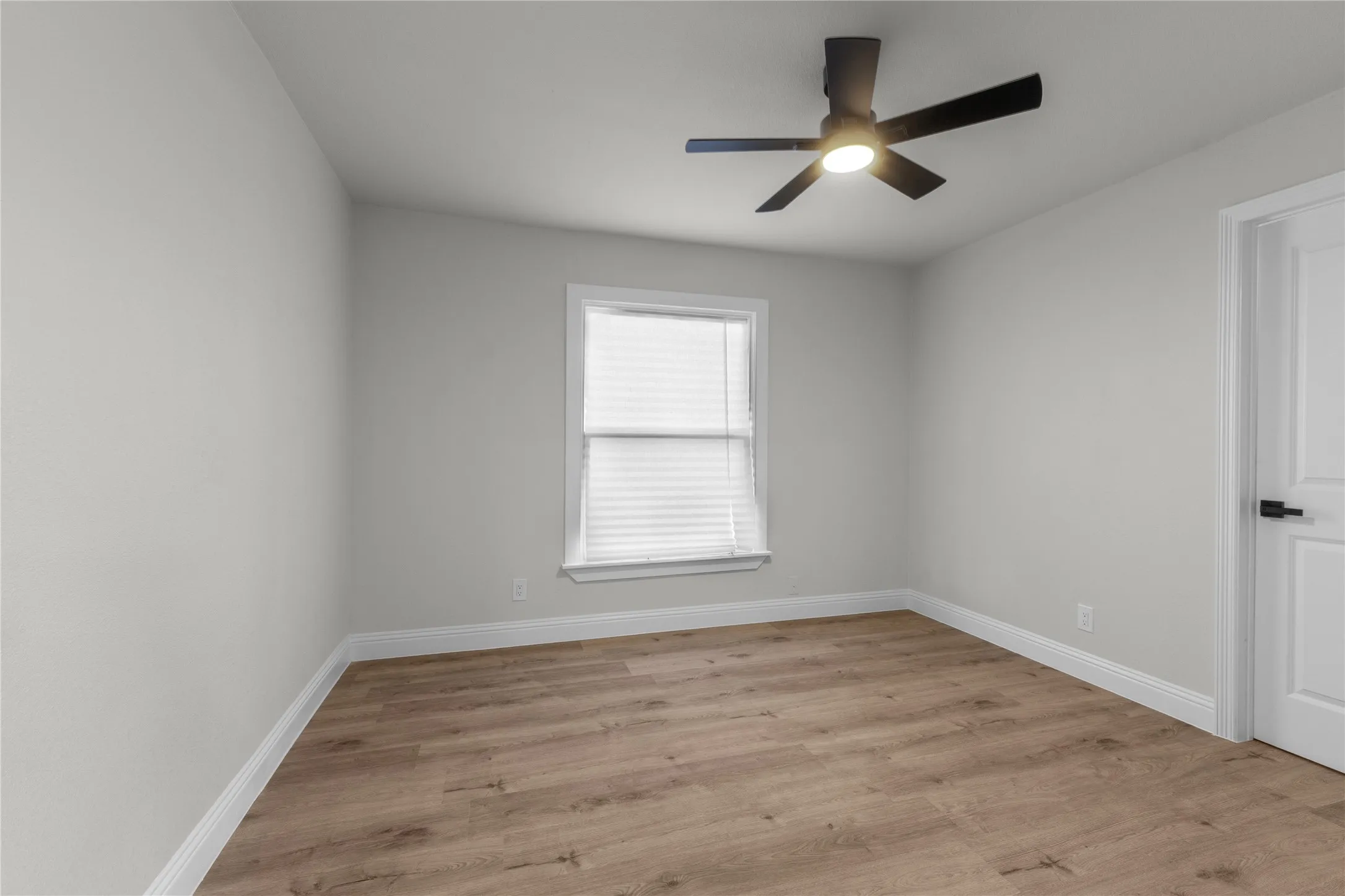 Wood-finish flooring, white walls, single window with blinds, ceiling fan with integrated lighting, and a white paneled door with dark hardware