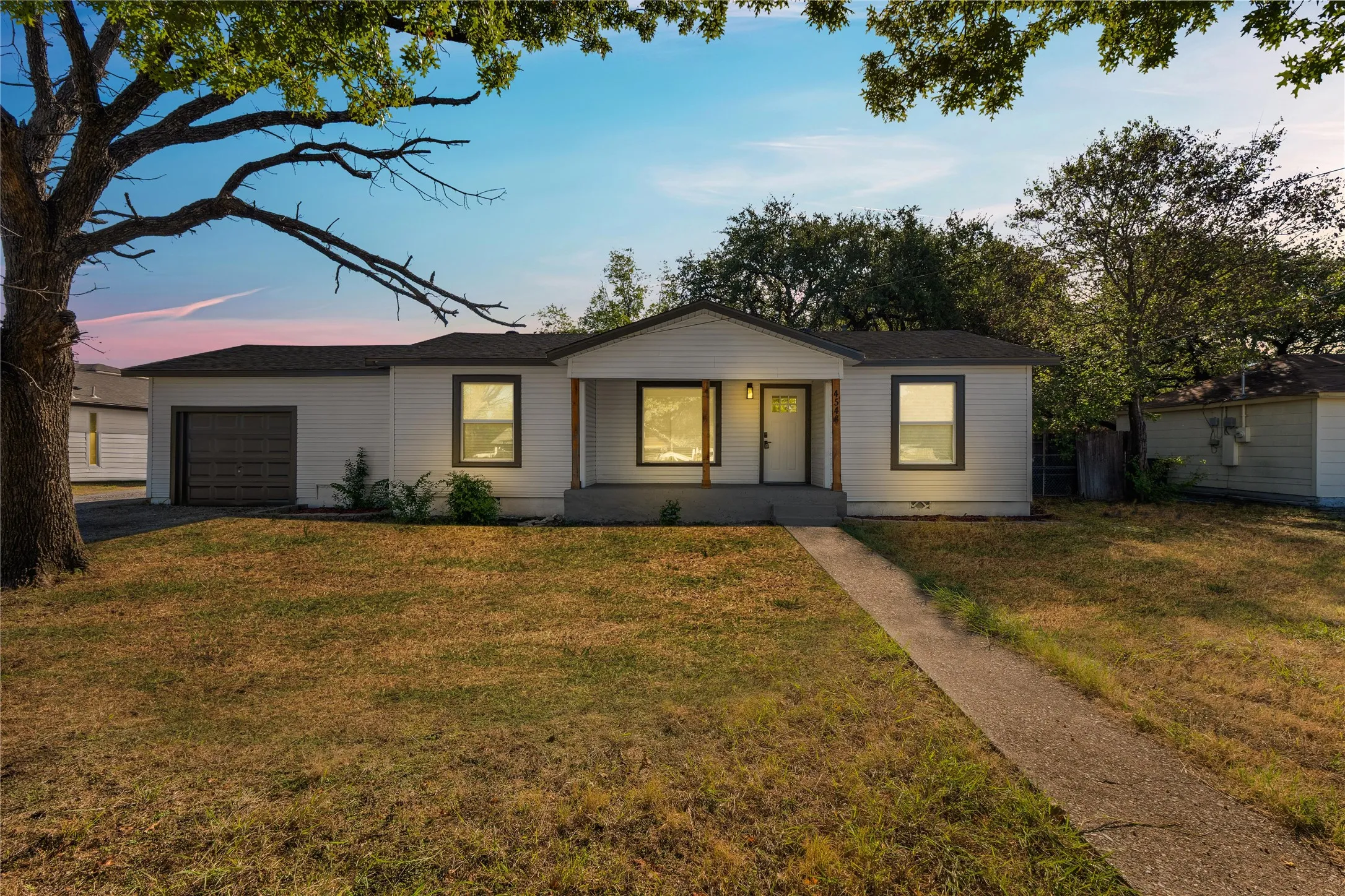 Modern single-story residence featuring a clean geometric facade with board-and-batten siding, expansive front-facing windows with warm interior lighting, an attached garage with dark paneled door, and a mature tree canopy providing natural shade to the manicured lawn and established landscaping