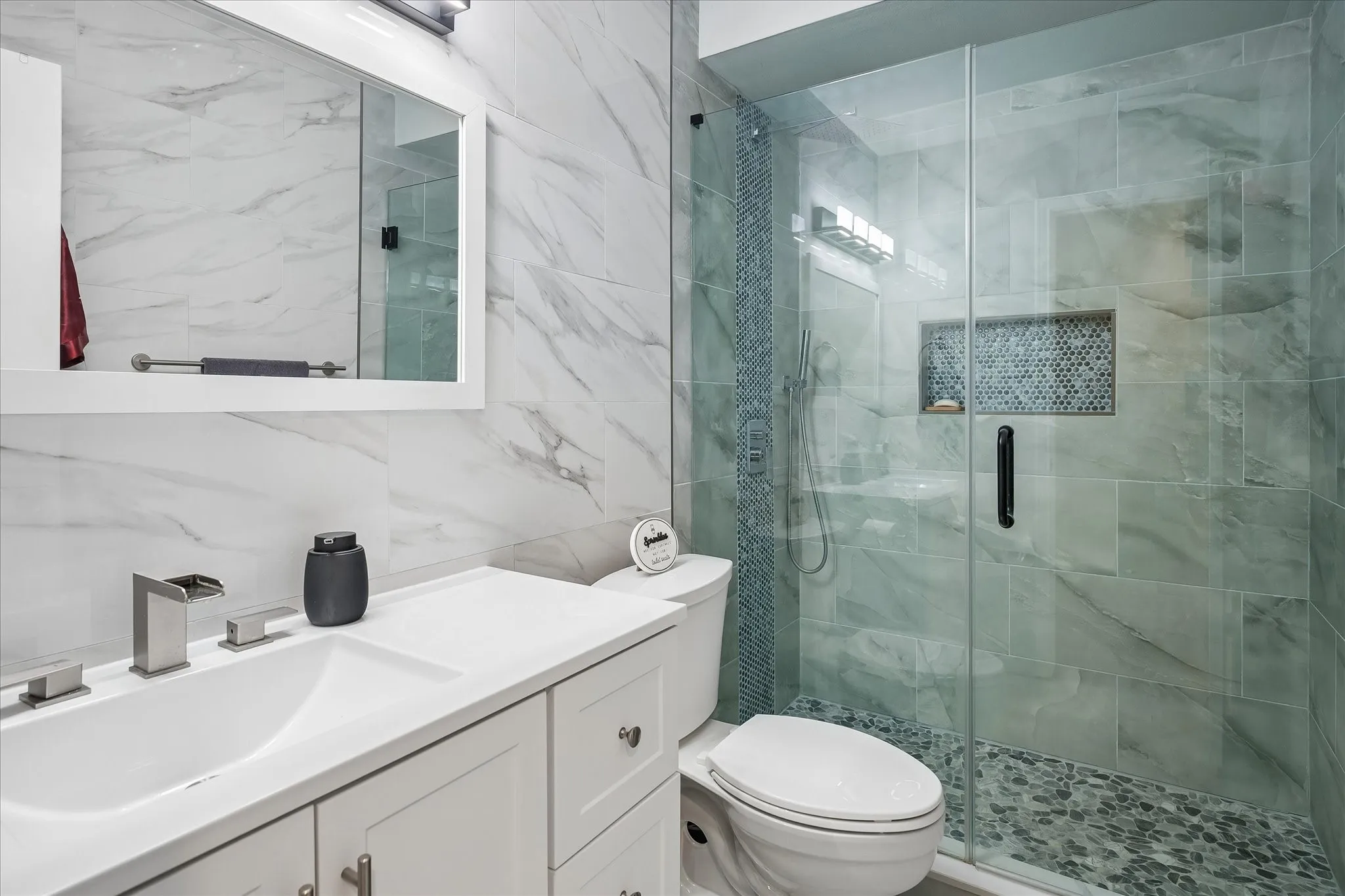 Bathroom featuring a white vanity cabinet with an integrated sink, wall-mounted mirror, glass-enclosed walk-in shower with stone tile flooring, and a built-in recessed wall niche