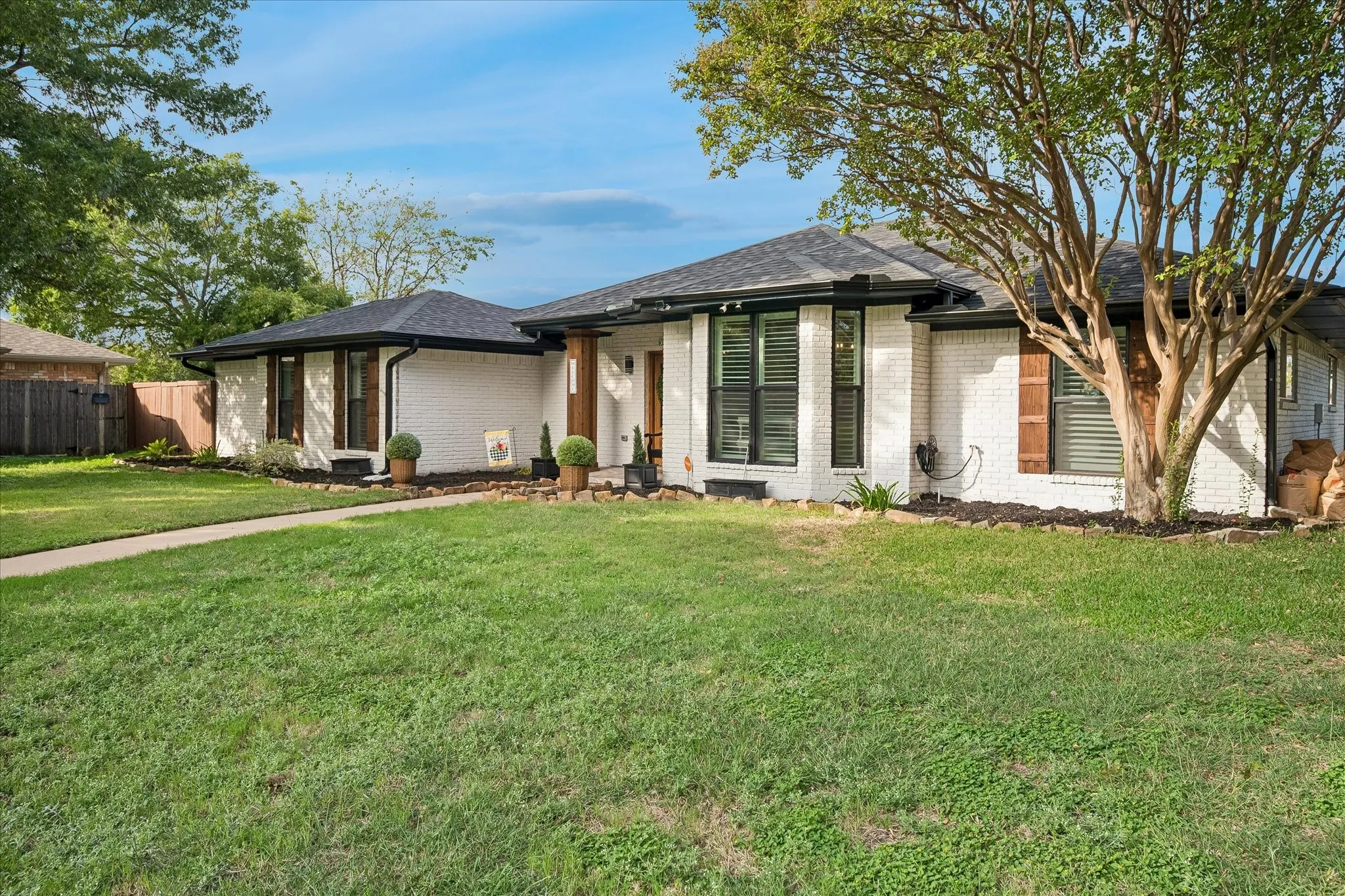 Single-story residence featuring white brick exterior, dark window trim, dark shingle roof, wood-tone column accent, and manicured lawn